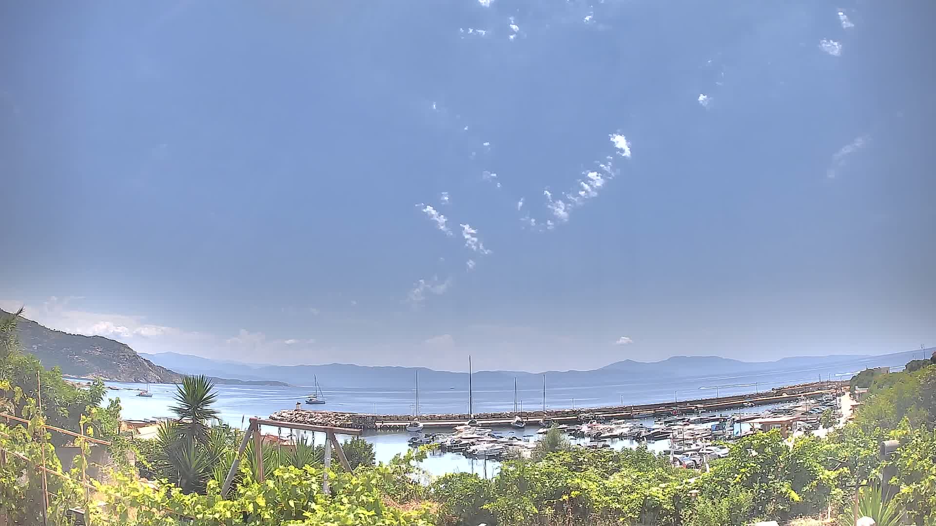 A marina filled with boats is seen from a hillside overlooking a calm, clear blue sea under a mostly sunny sky with scattered clouds.