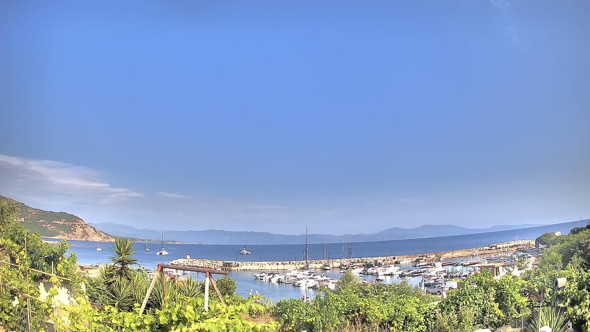 A marina filled with boats is nestled between green hills and a calm, blue sea under a mostly clear sky.