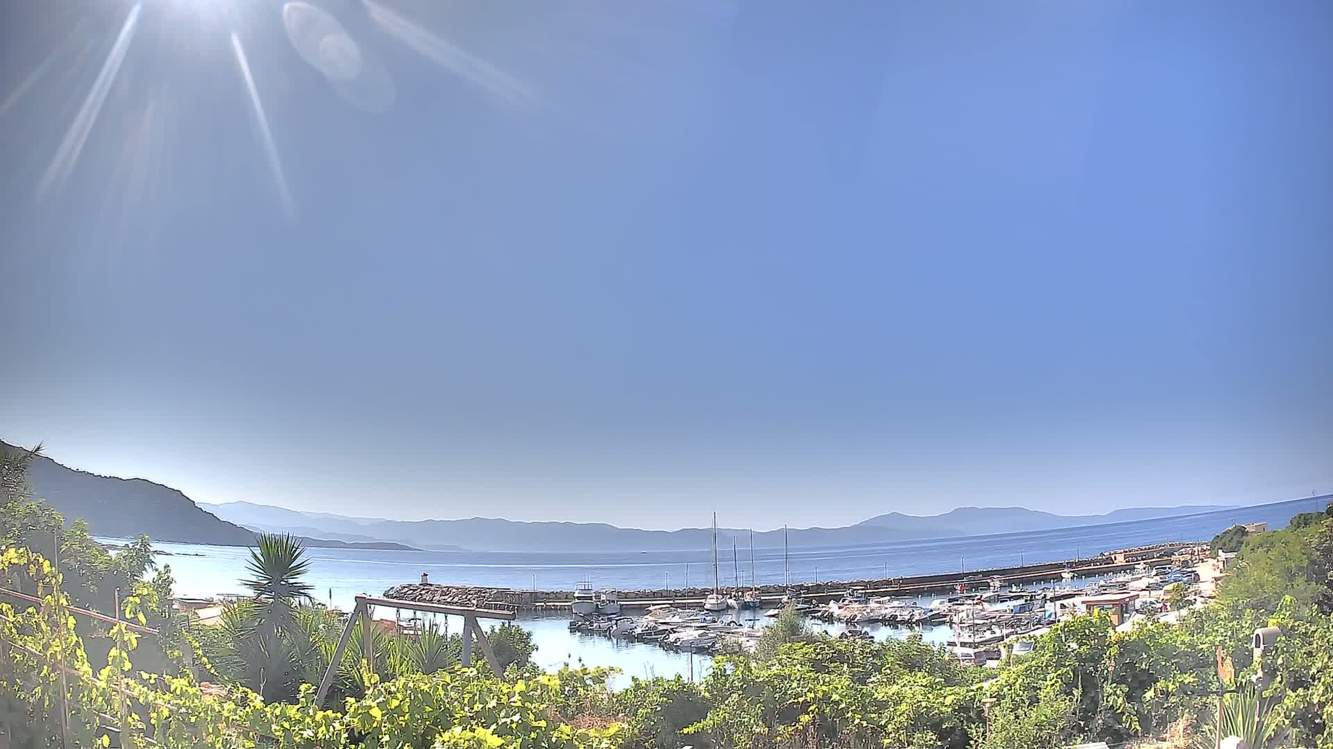 A sunny day overlooks a calm harbor filled with boats, nestled between green hills and a clear blue sea under a bright sky.