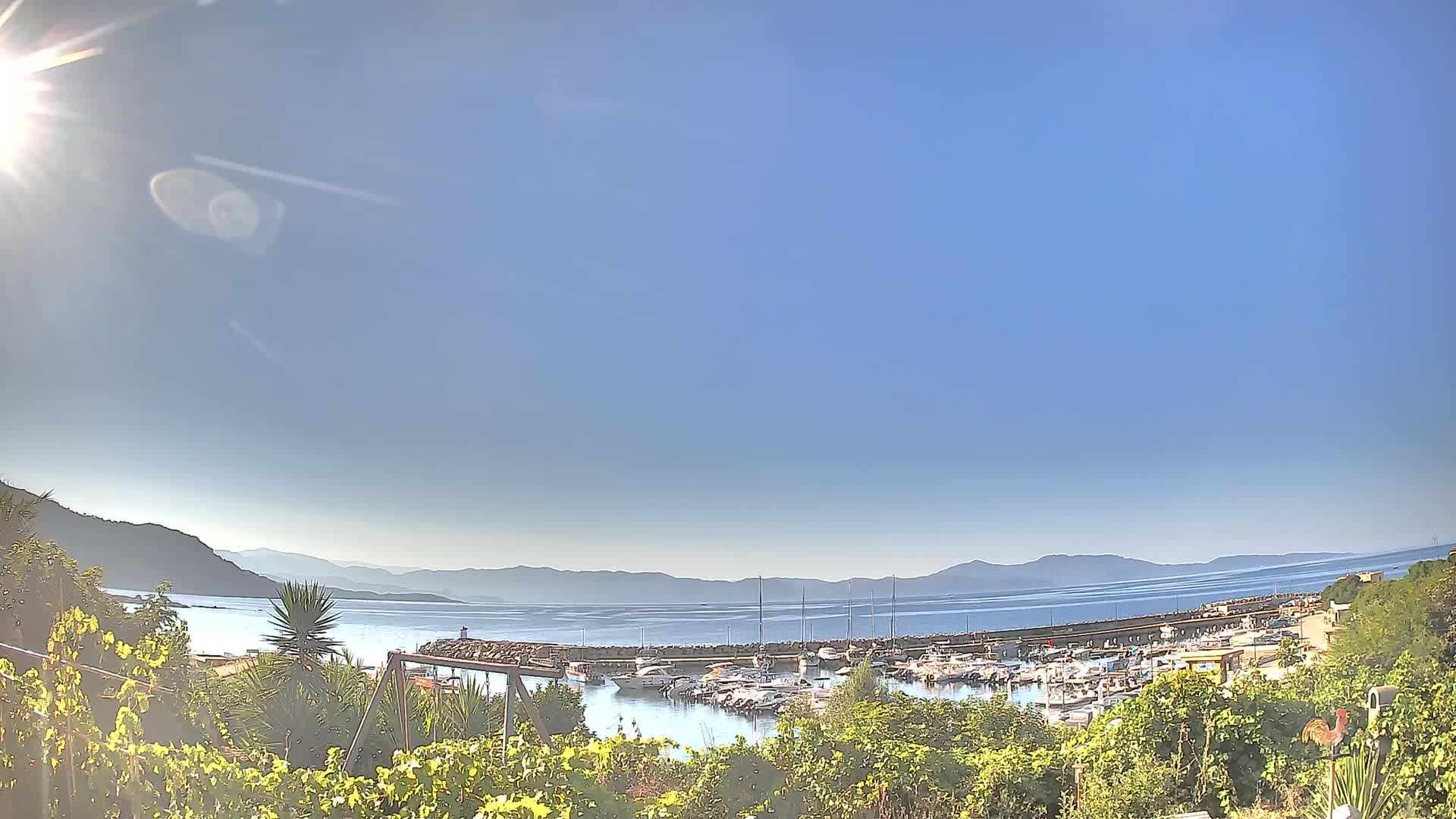 A sunny day reveals a harbor filled with boats, nestled between hills and a calm sea under a clear blue sky.