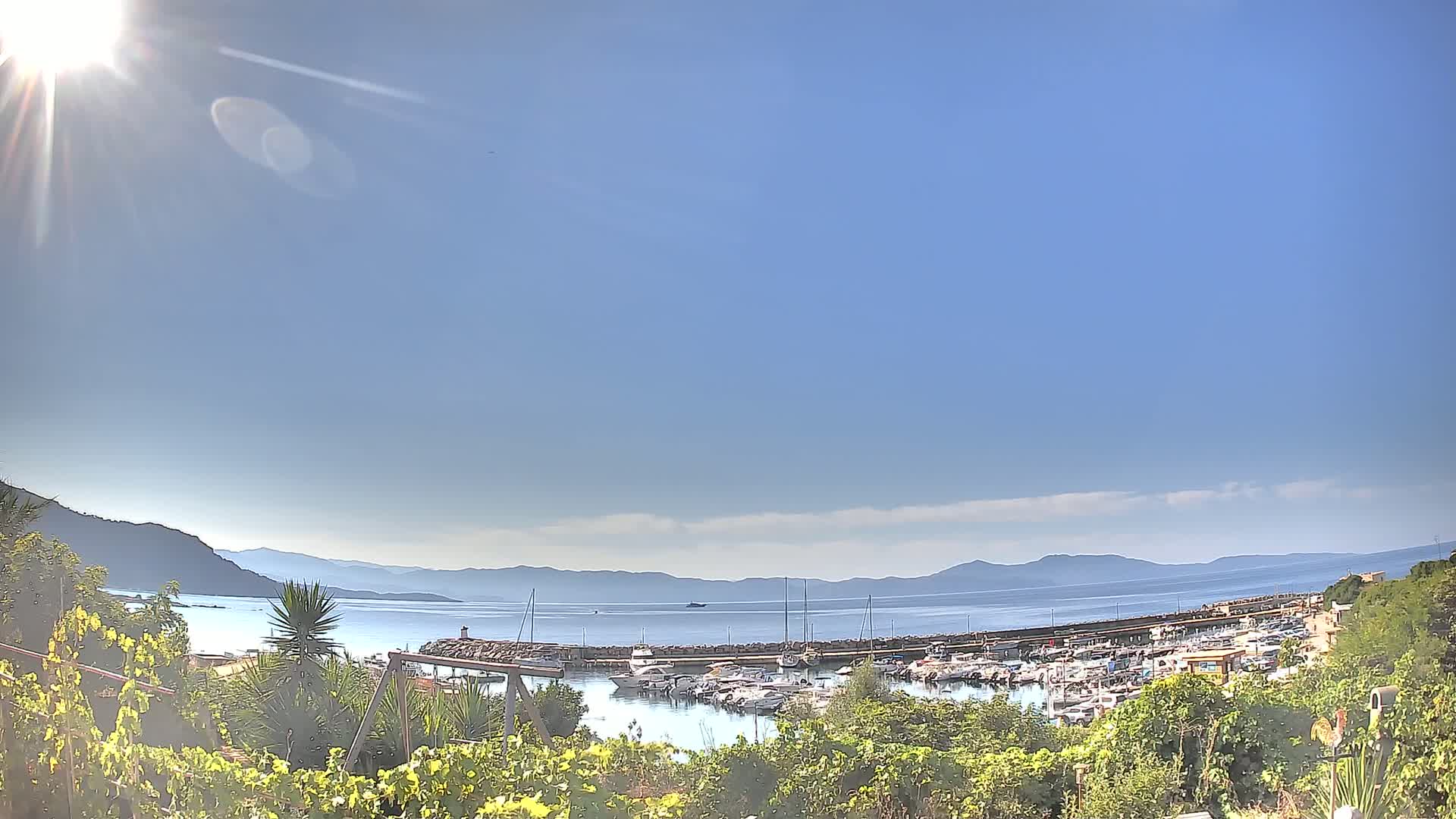 A sunny day reveals a marina filled with boats, nestled between a verdant hillside and a calm sea stretching to distant mountains.