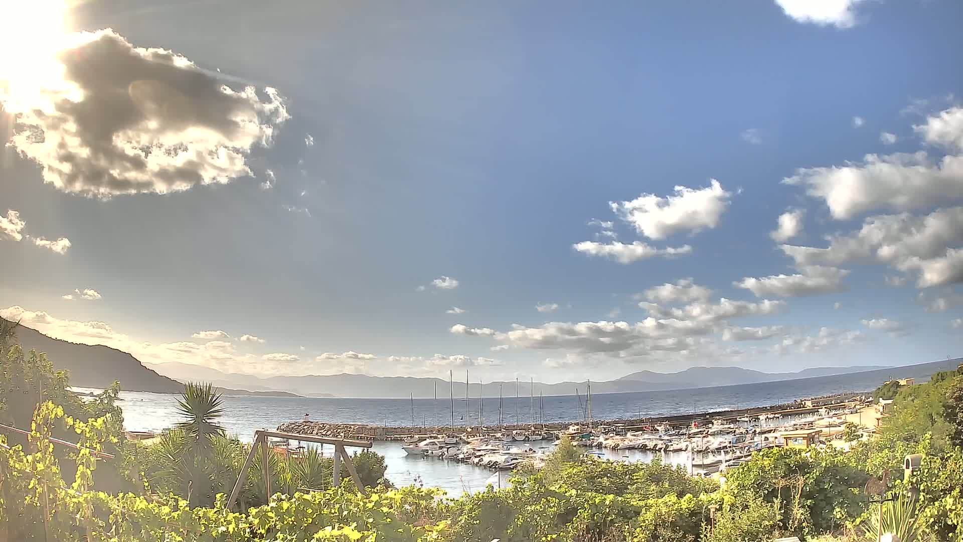 A partly sunny day reveals a marina filled with boats, nestled between green vegetation and a calm sea with distant mountains under a partly cloudy sky.