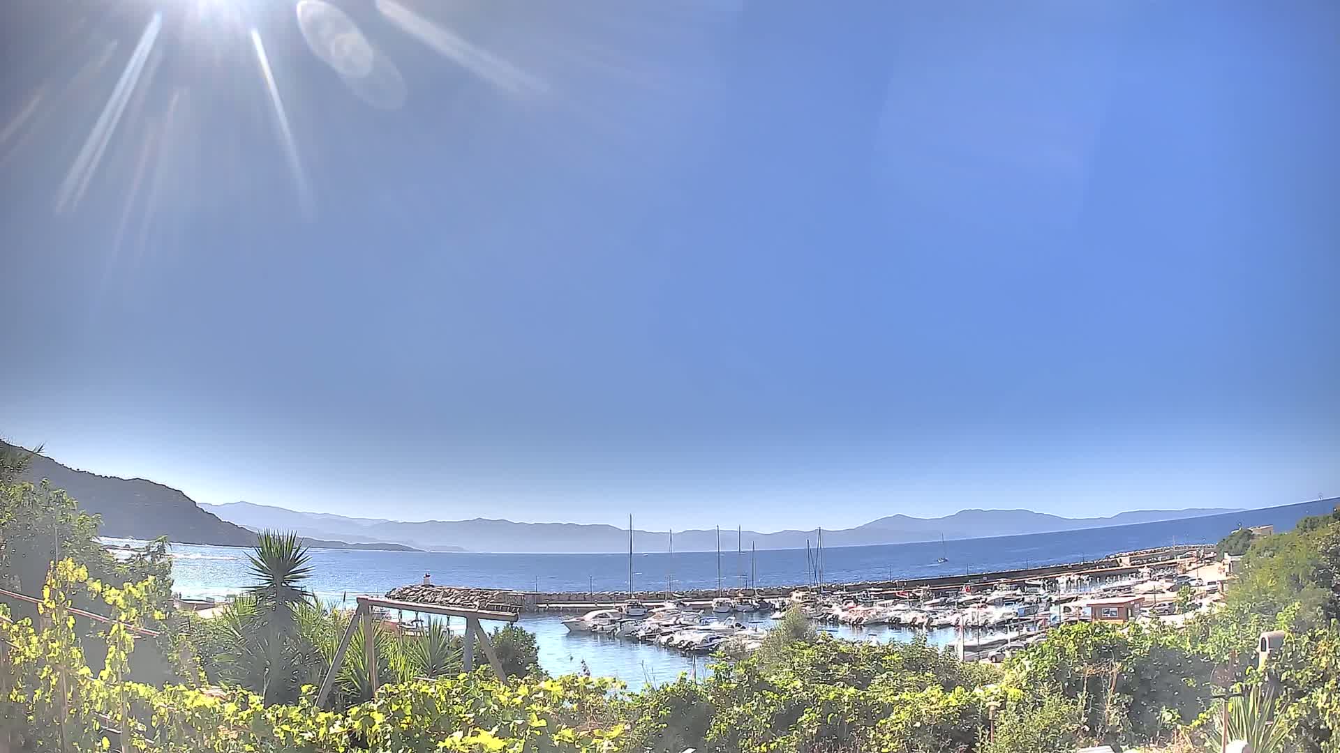 A sunny day reveals a marina filled with boats, nestled between a green hillside and a calm, blue sea with distant mountains under a clear sky.