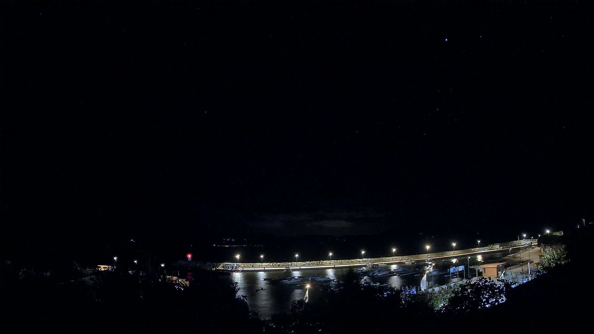 A sunny day overlooking a harbor filled with boats, nestled between a verdant hillside and a calm sea with distant mountains.