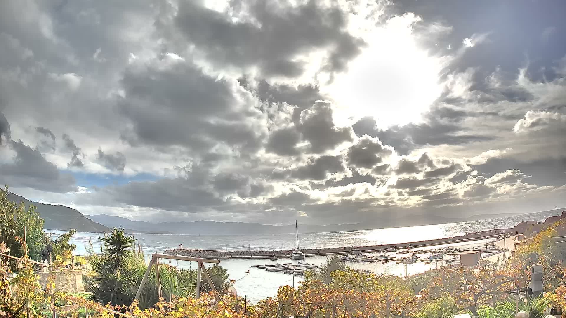 Under a dramatic sky of heavy clouds with a brilliant sun breaking through, a coastal harbor filled with numerous boats is visible, framed by autumnal foliage and distant mountains, with sunlight intensely reflecting on the choppy water.
