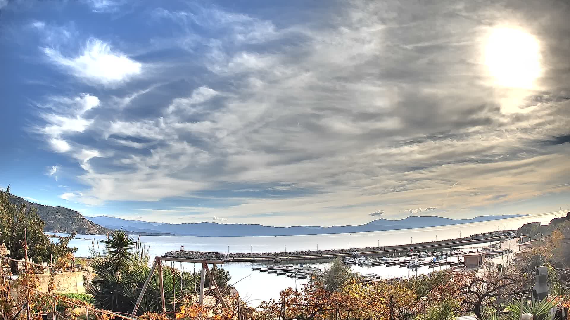Under a dramatic sky of heavy clouds with a brilliant sun breaking through, a coastal harbor filled with numerous boats is visible, framed by autumnal foliage and distant mountains, with sunlight intensely reflecting on the choppy water.