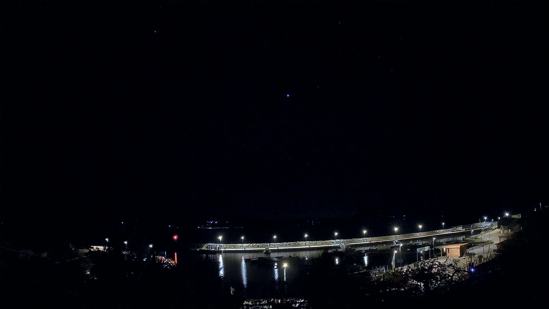 Under a dramatic sky of heavy clouds with a brilliant sun breaking through, a coastal harbor filled with numerous boats is visible, framed by autumnal foliage and distant mountains, with sunlight intensely reflecting on the choppy water.