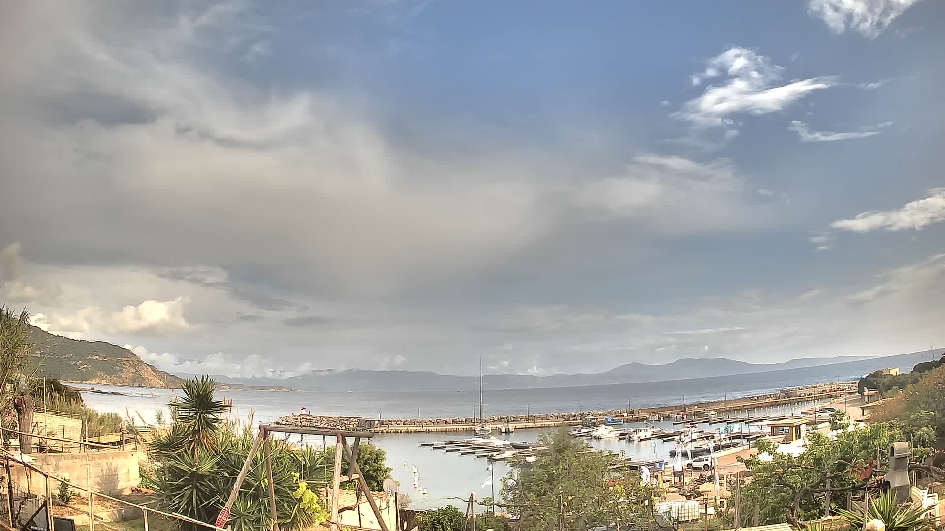 Under a dramatic sky of heavy clouds with a brilliant sun breaking through, a coastal harbor filled with numerous boats is visible, framed by autumnal foliage and distant mountains, with sunlight intensely reflecting on the choppy water.