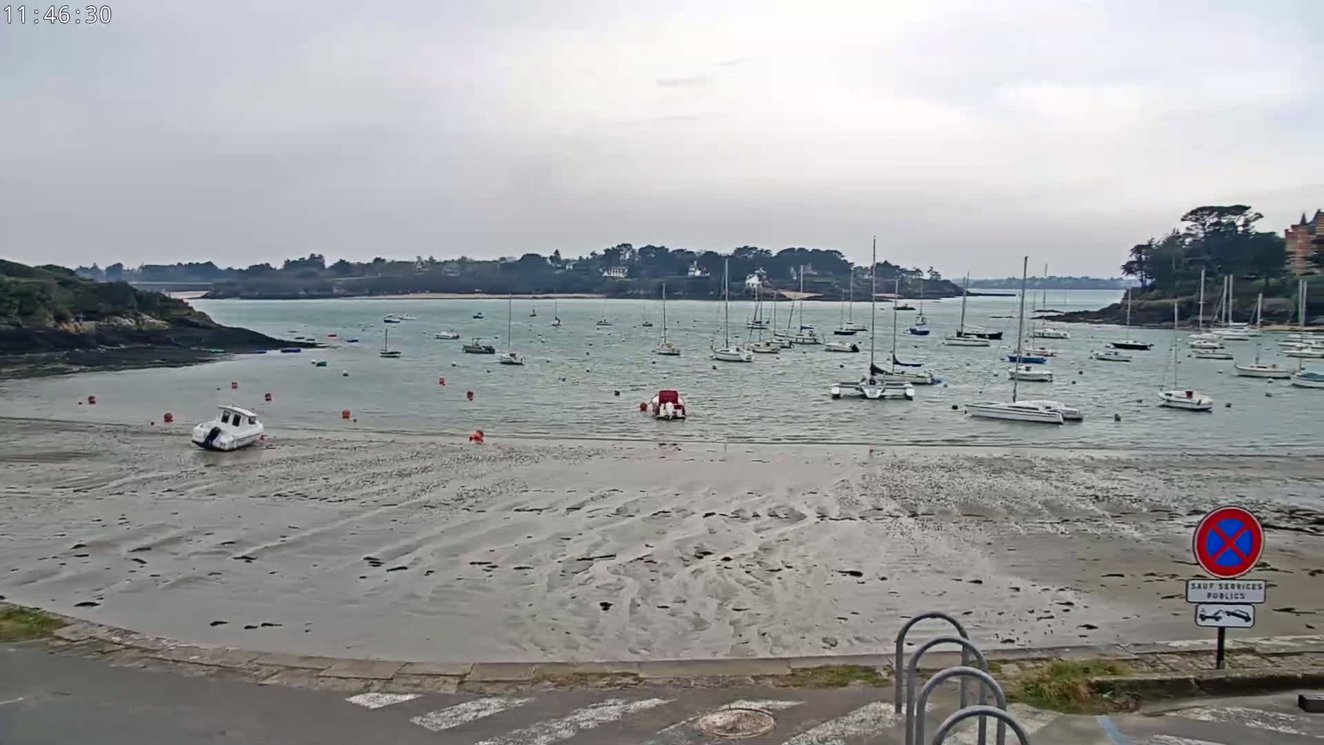 A calm, overcast day reveals a harbor filled with numerous sailboats and small motorboats at low tide, a sandy beach exposed in the foreground.