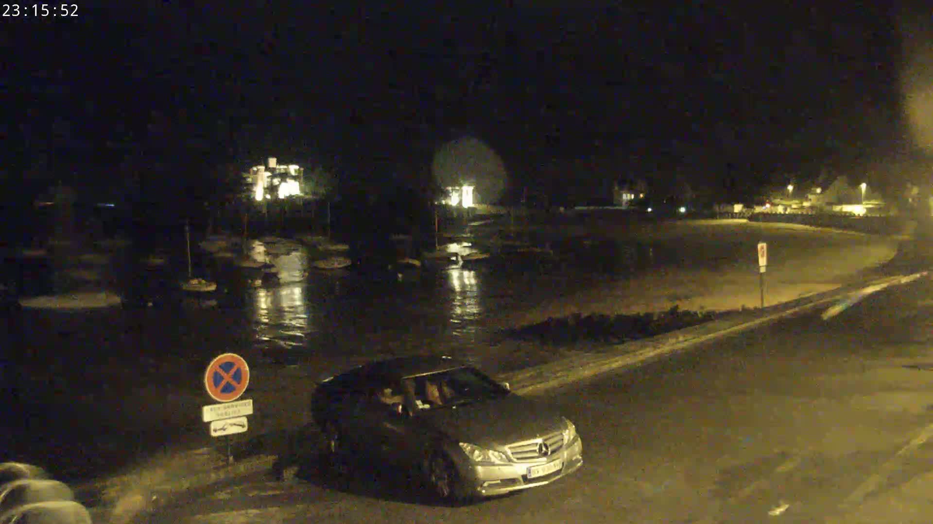 A silver car is parked near a nighttime harbor with several boats, under a dark sky.