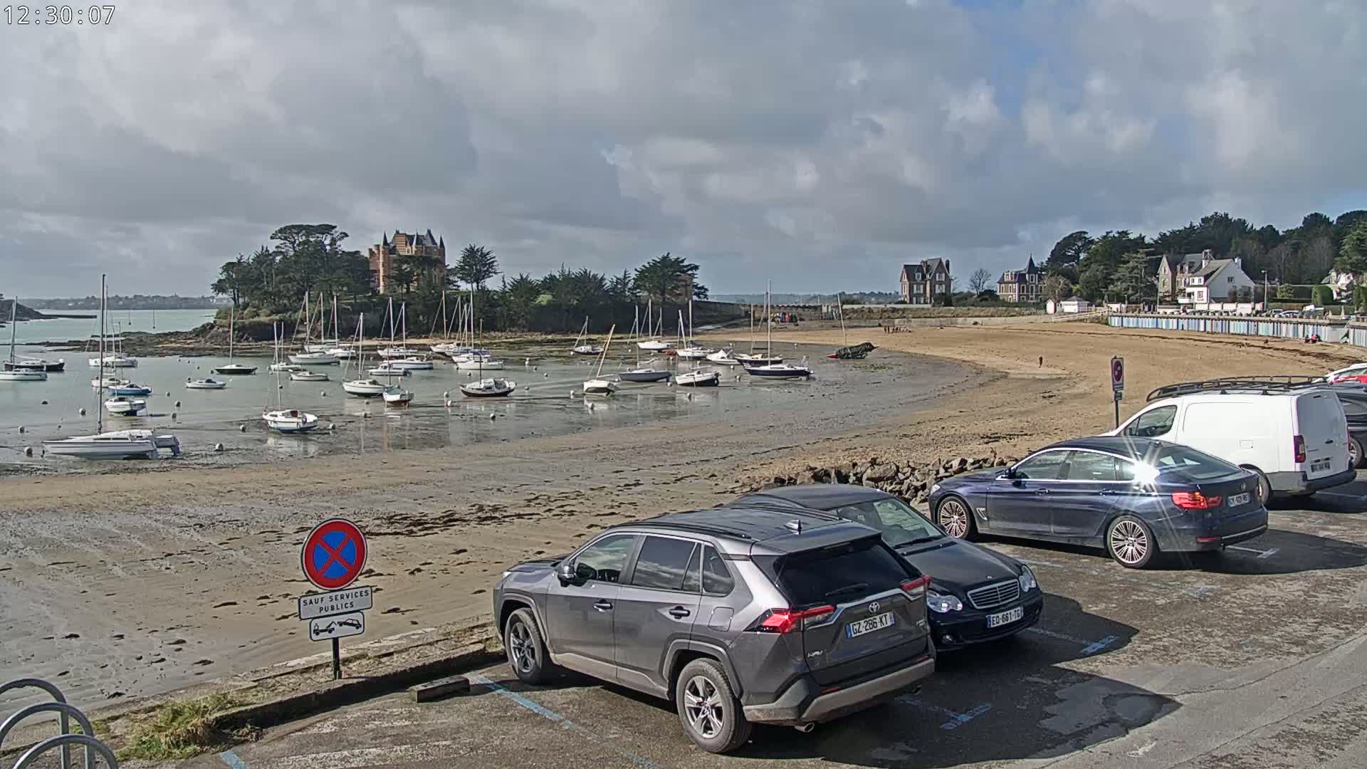 A sandy beach with numerous sailboats in the shallow water near a stone chateau and buildings, under a partly cloudy sky.