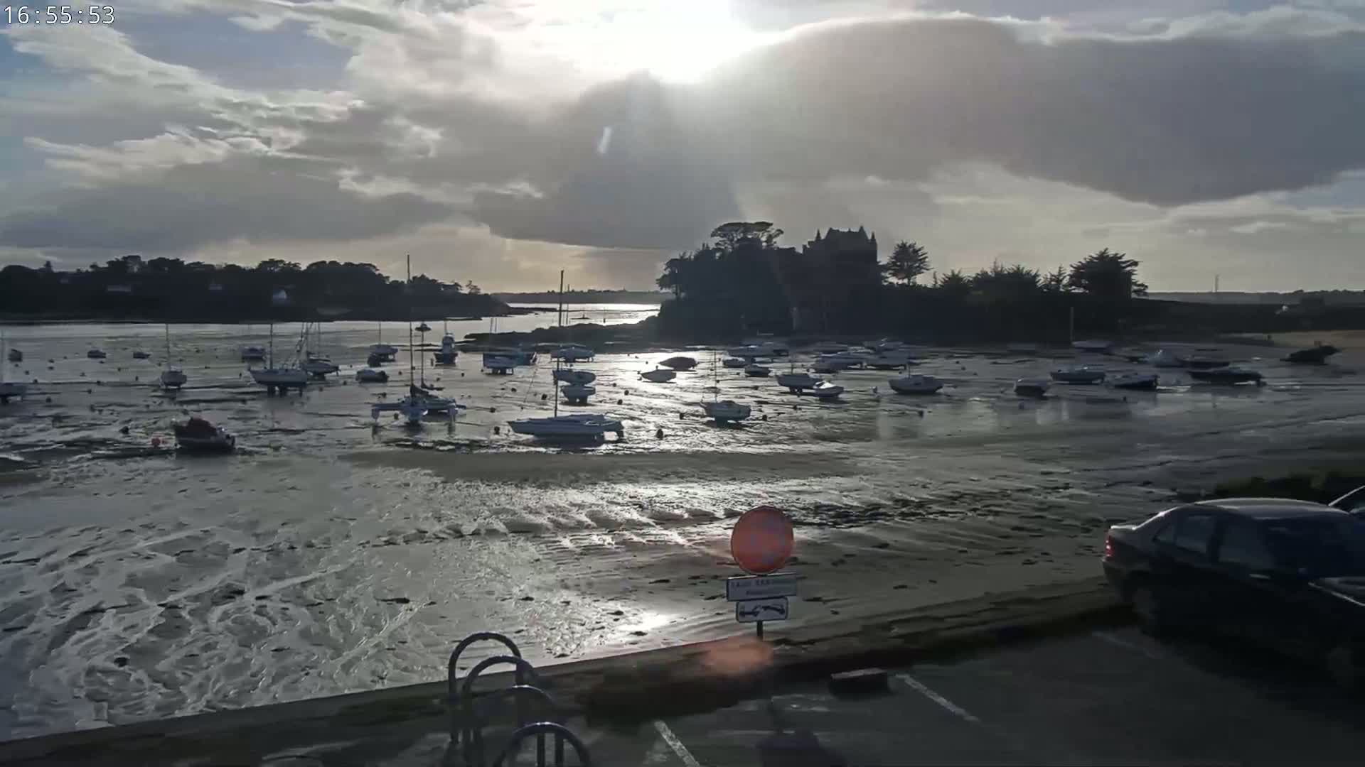 Numerous sailboats sit aground on a tidal flat near a stone building, under a partly cloudy sky with the sun shining.