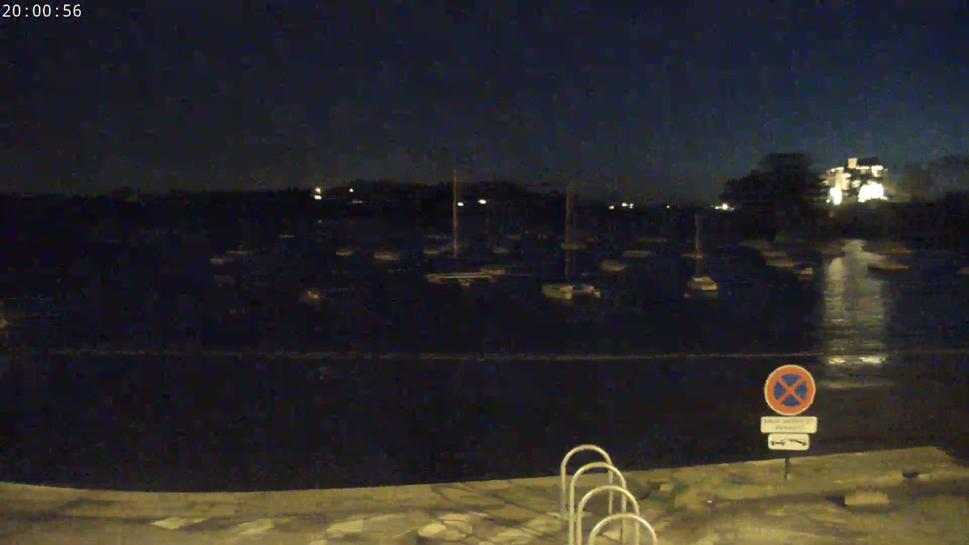 A nighttime view of a calm body of water with numerous boats moored, a no parking sign, and some metal ladders visible on the shore.