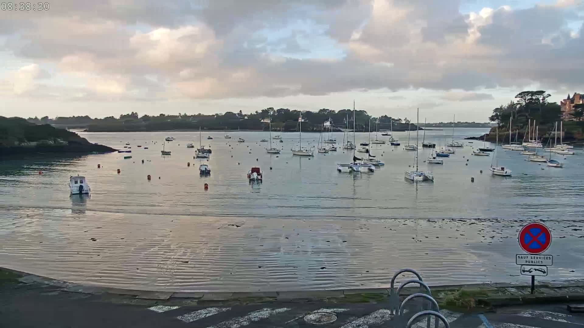 Many sailboats are moored in a calm bay under a partly cloudy sky.