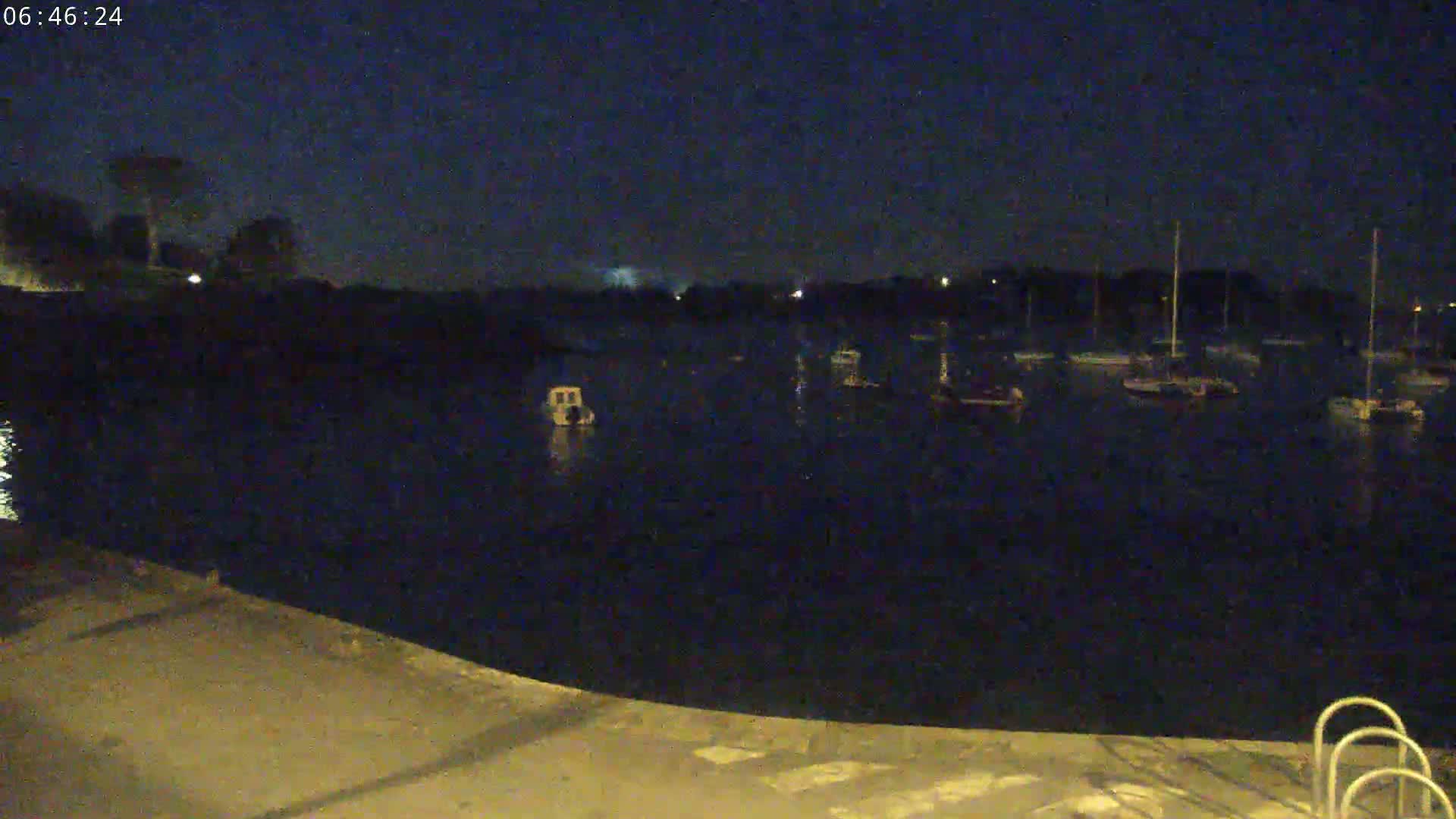 A nighttime view of a calm body of water with several boats moored, partially illuminated under a dark sky.
