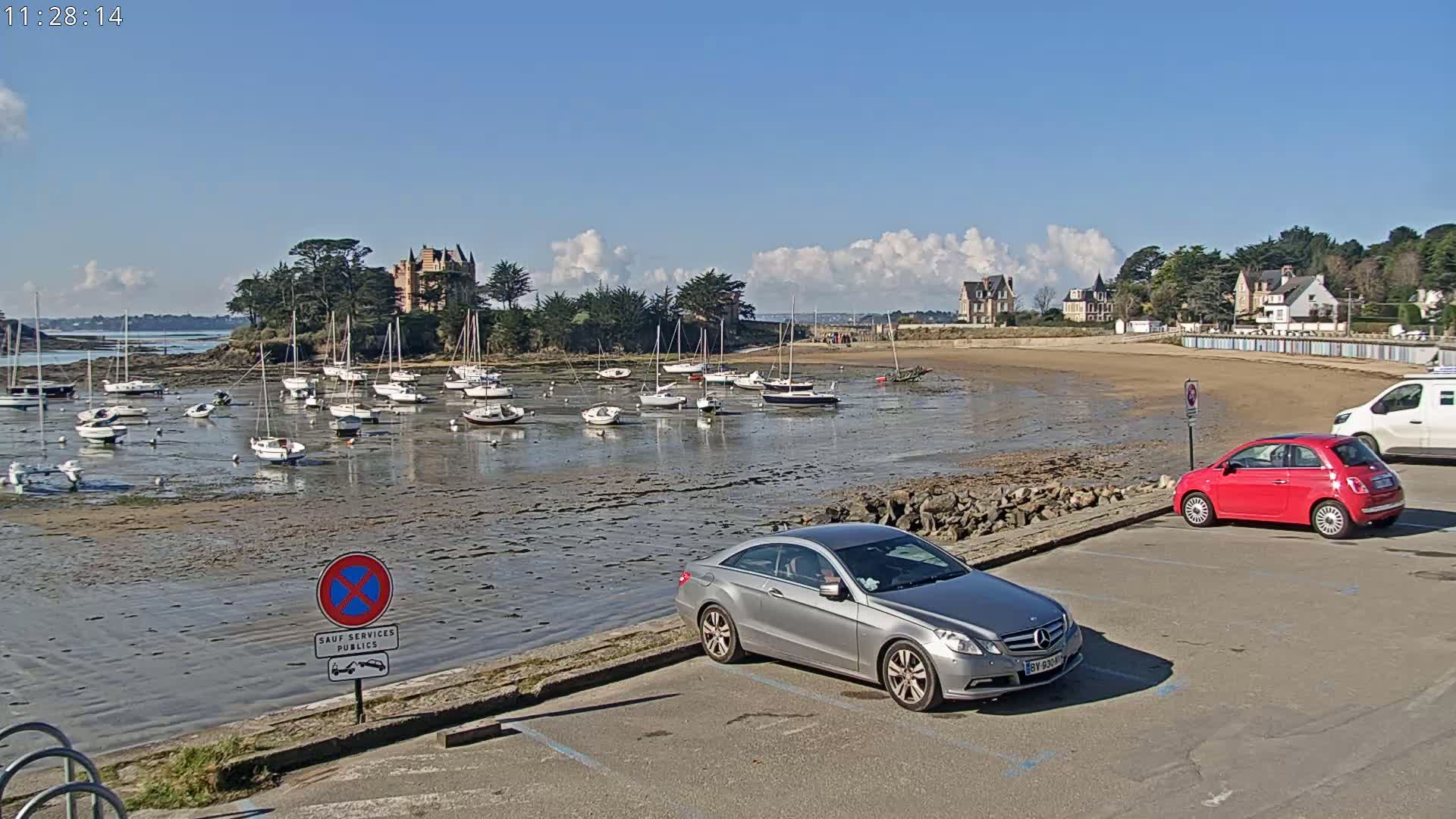 A harbor with numerous sailboats at low tide, under a partly cloudy sky, is viewed from a parking lot with cars.