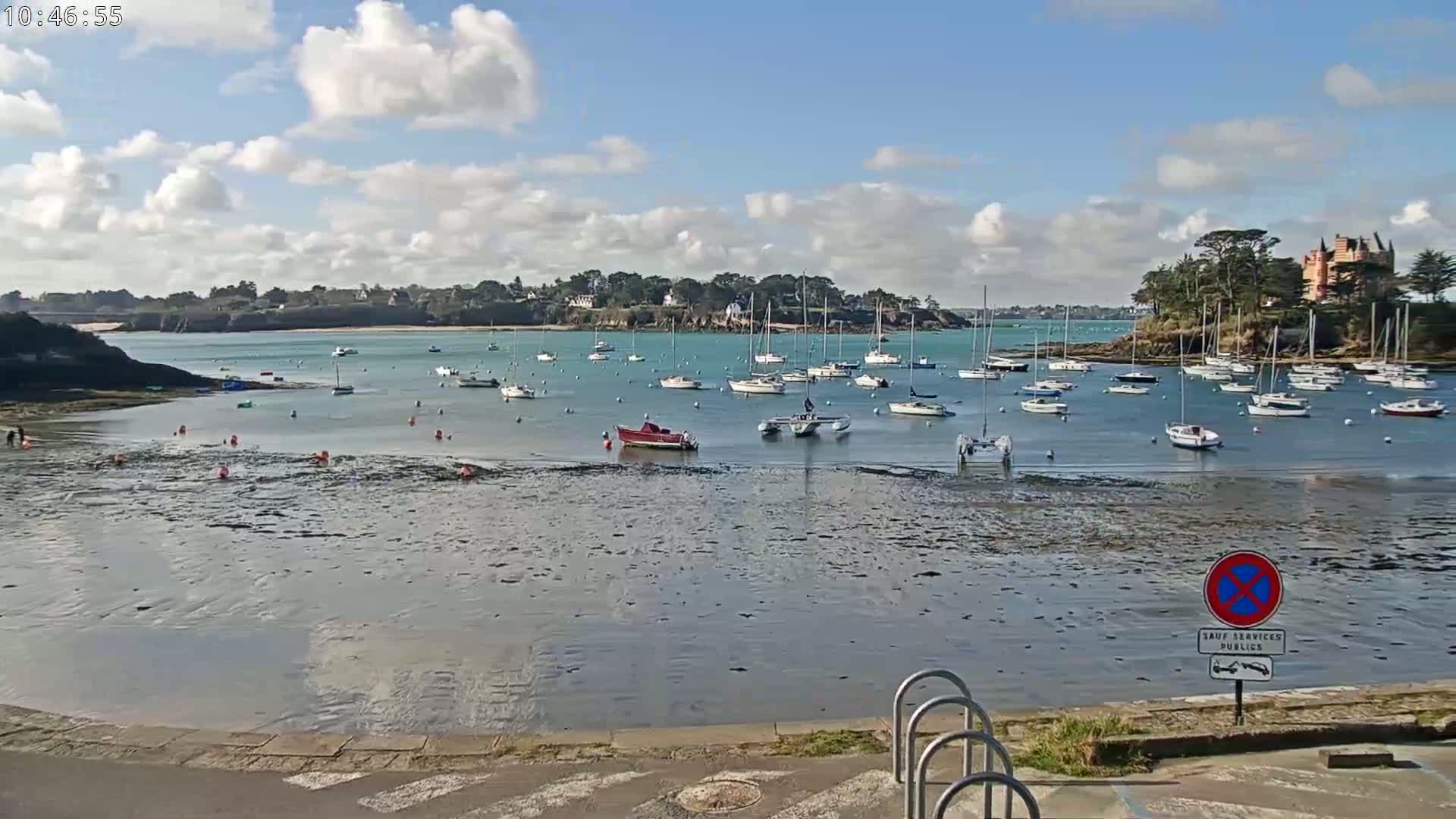 A calm bay with numerous sailboats and a few motorboats is partially exposed at low tide under a partly cloudy sky.