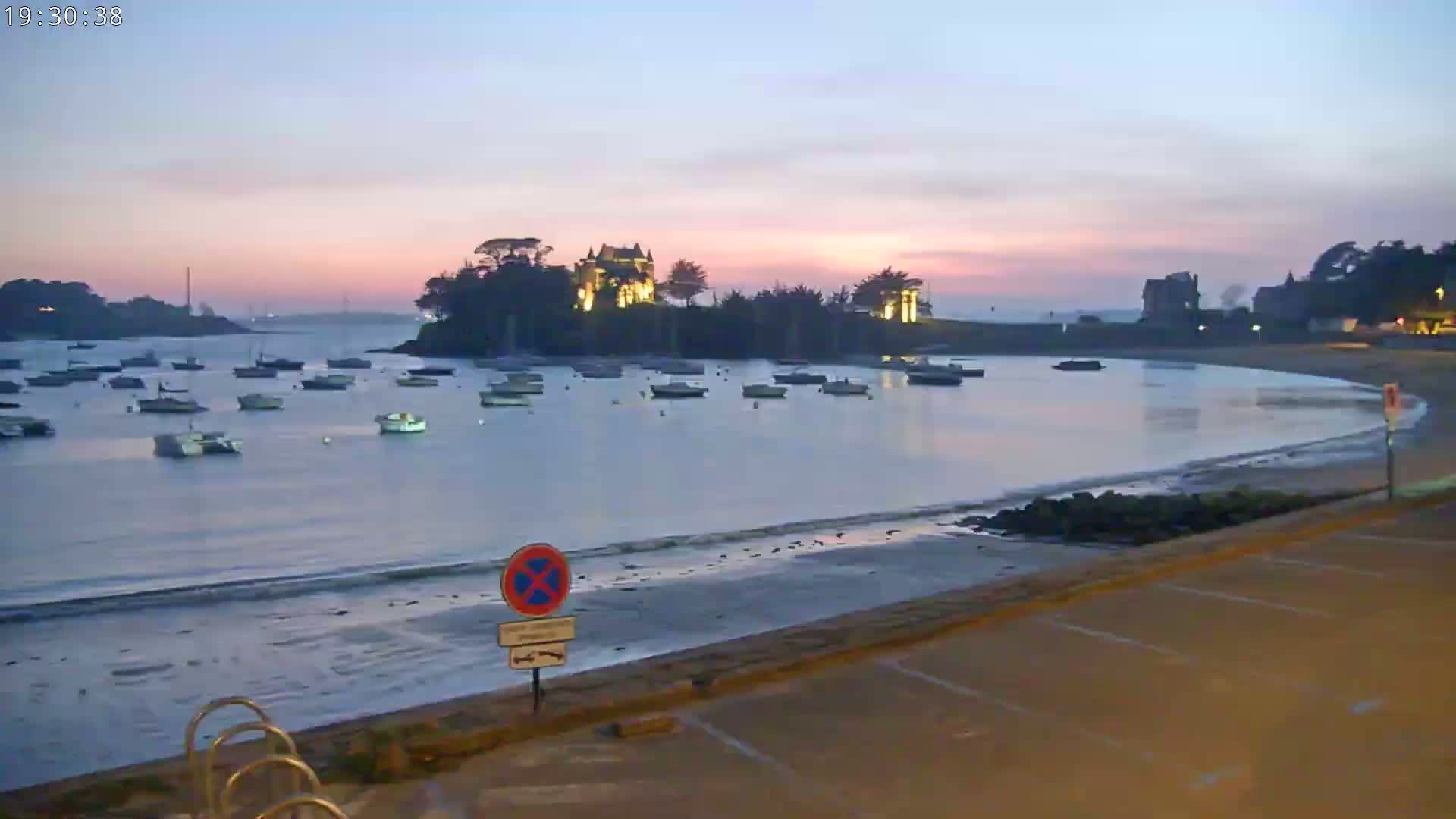 A calm bay at sunset, with numerous small boats moored in the water, a sandy beach, and a lit-up building on a small island in the background.