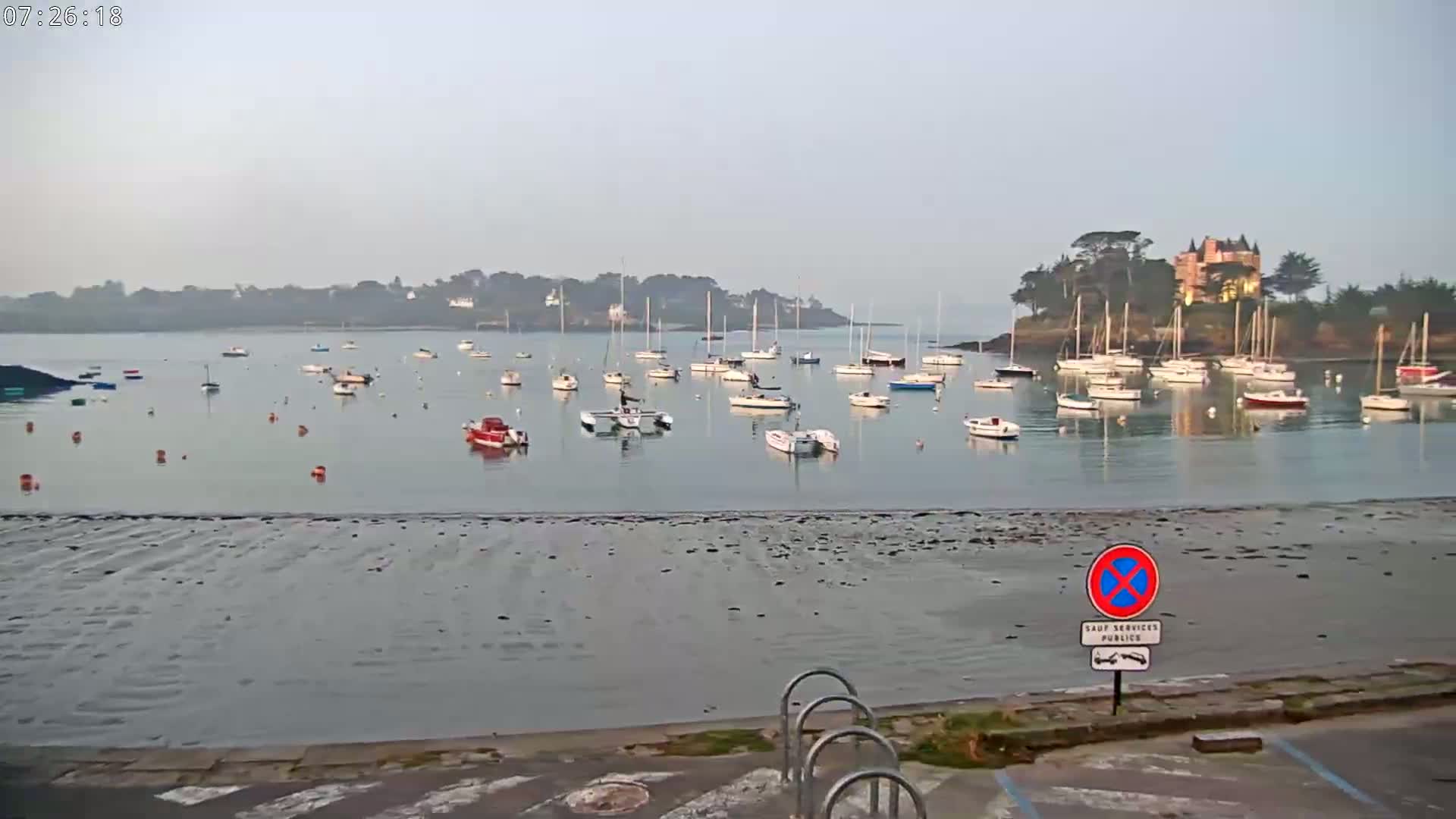 Many sailboats are moored in a calm bay on a hazy day, with a large house visible on a nearby island.