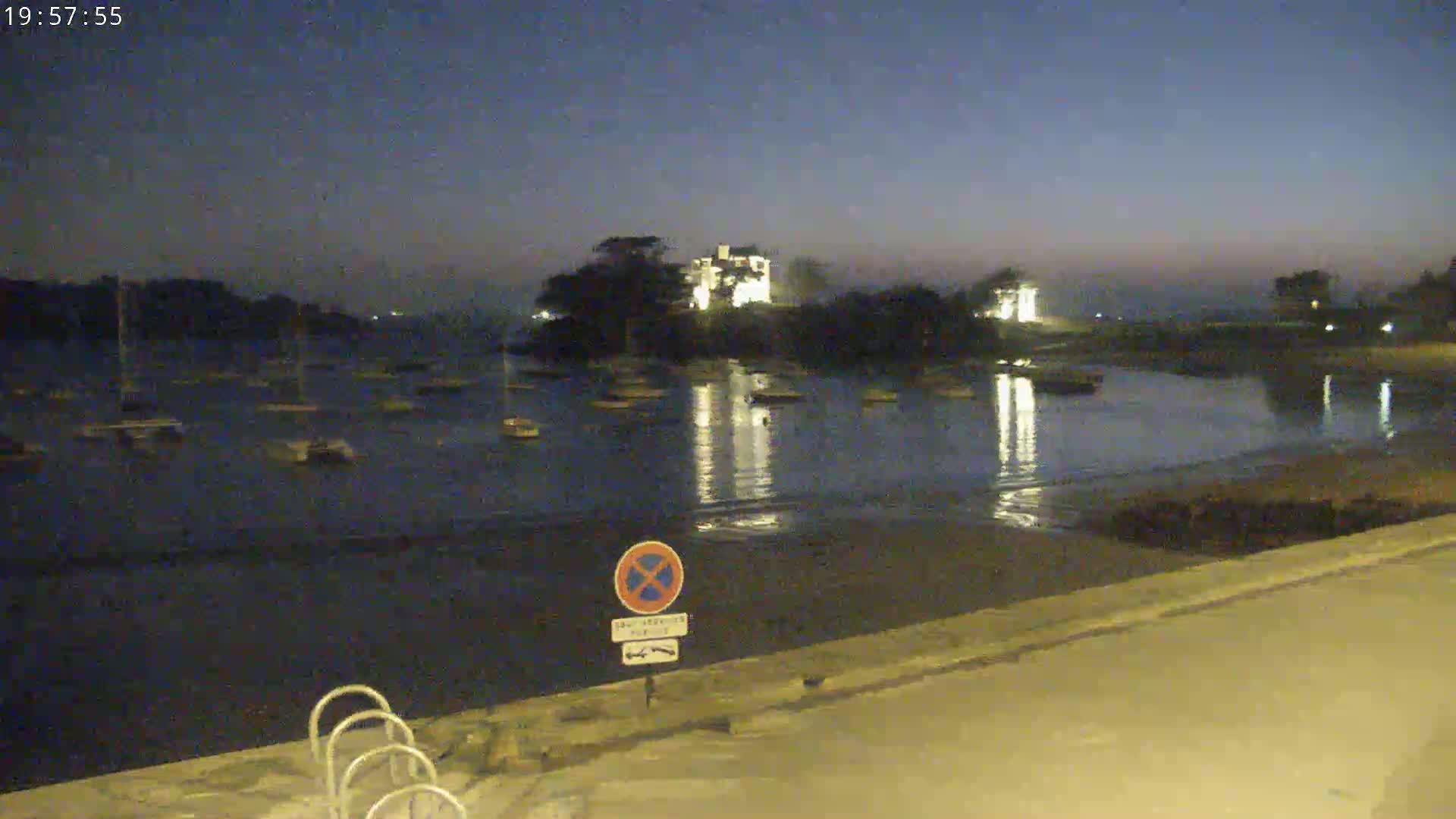 A calm body of water at twilight is dotted with numerous small boats, reflecting lights from a building on a small island in the distance.