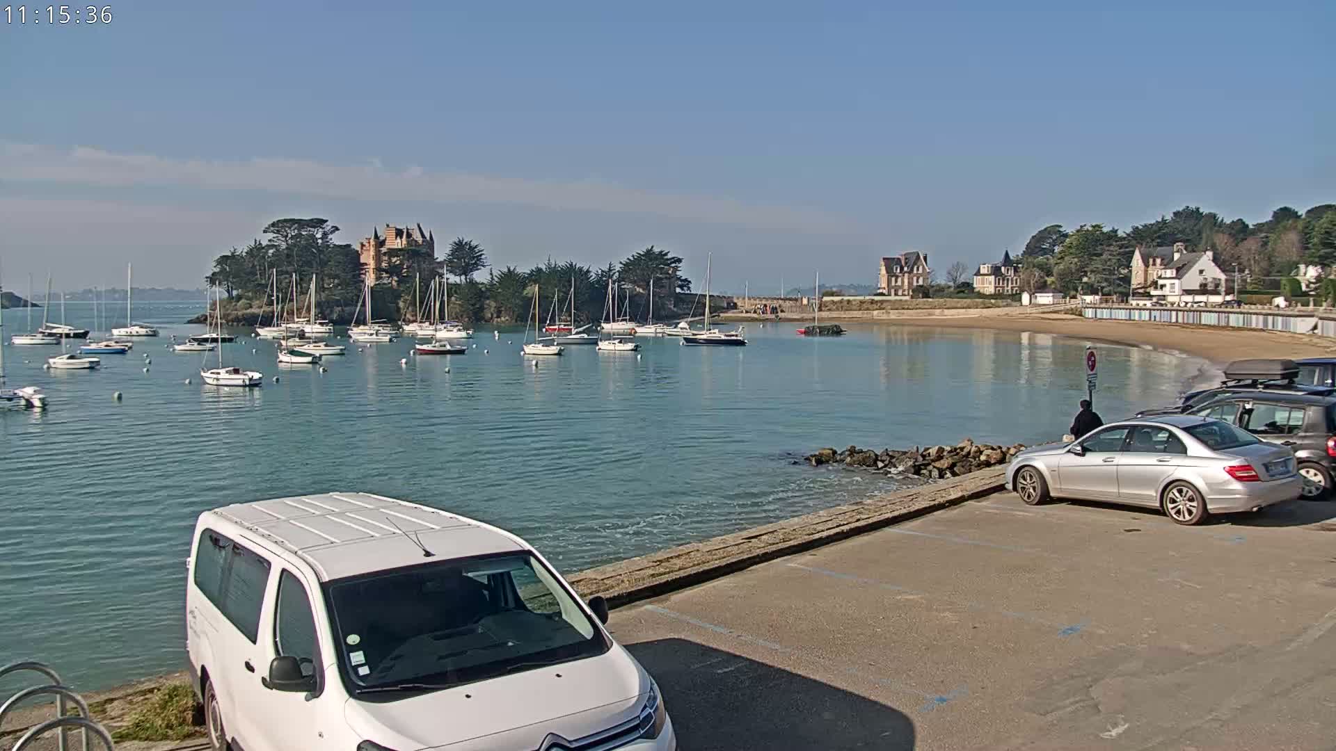 A calm bay with numerous sailboats, a small sandy beach, several buildings, and a parking lot with cars, all under a clear, sunny sky.