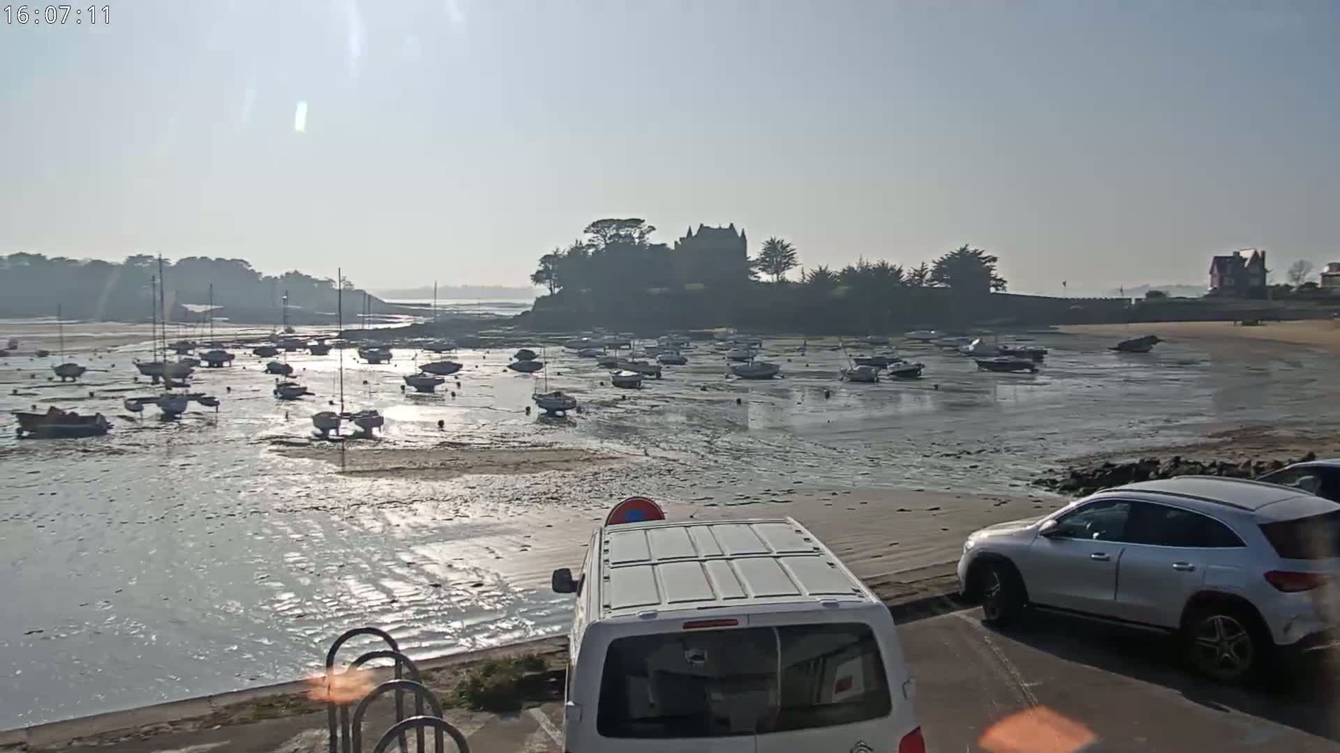 Numerous sailboats are grounded on a tidal flat near a shoreline with a building on a small island, under a hazy sunny sky.