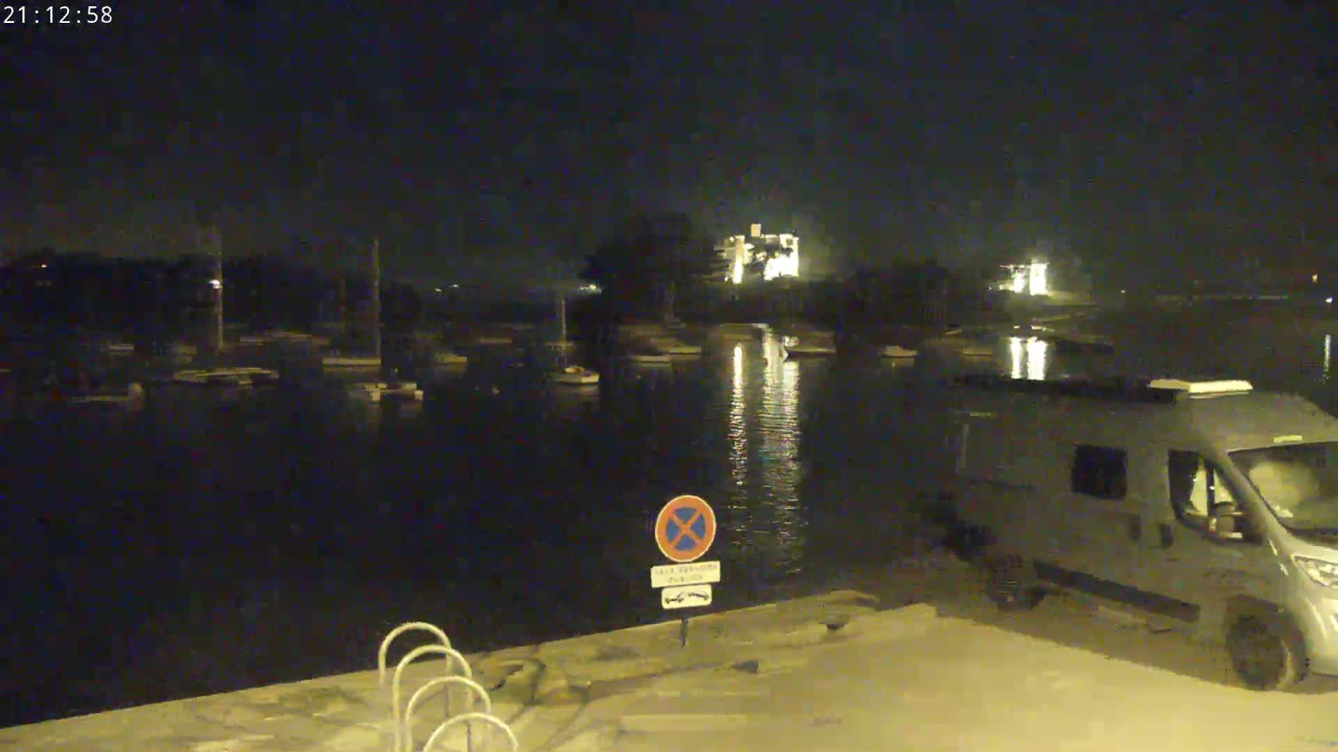 At night, a campervan is parked near a calm body of water with boats moored, illuminated by lights from buildings in the distance.