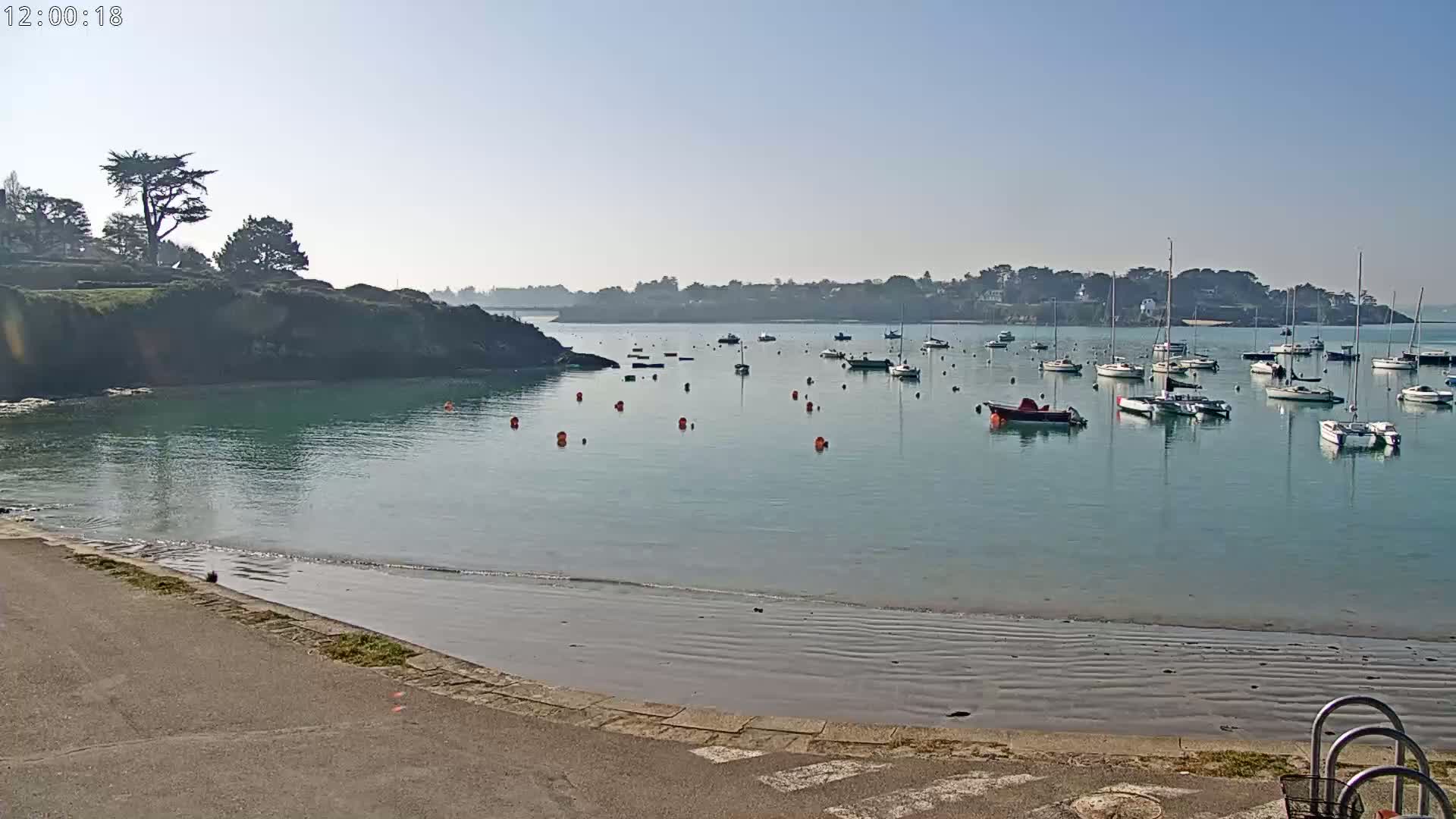 A calm, sunny day shows a tranquil bay with numerous sailboats and motorboats at anchor, a sandy beach, and a small rocky headland with greenery.