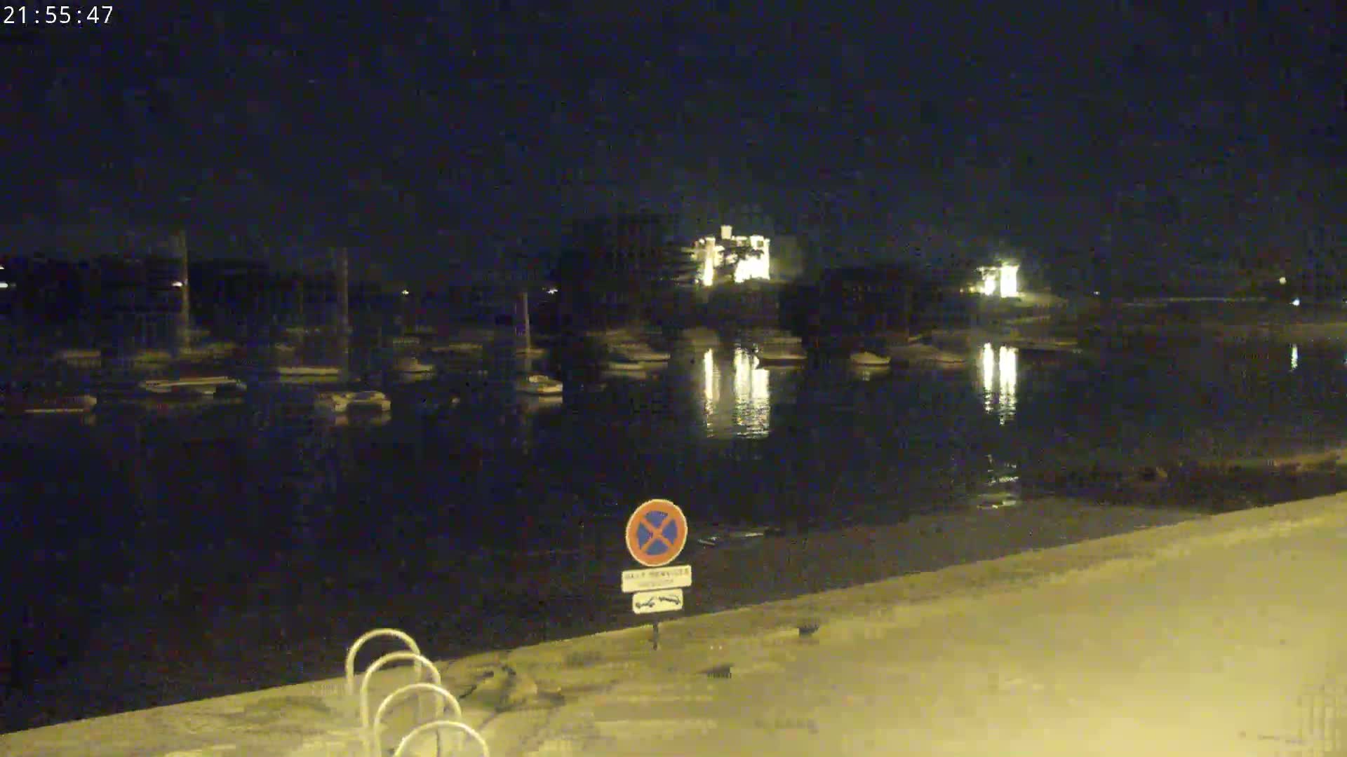 A nighttime view of a calm body of water reflecting lights from buildings and boats across the shore.