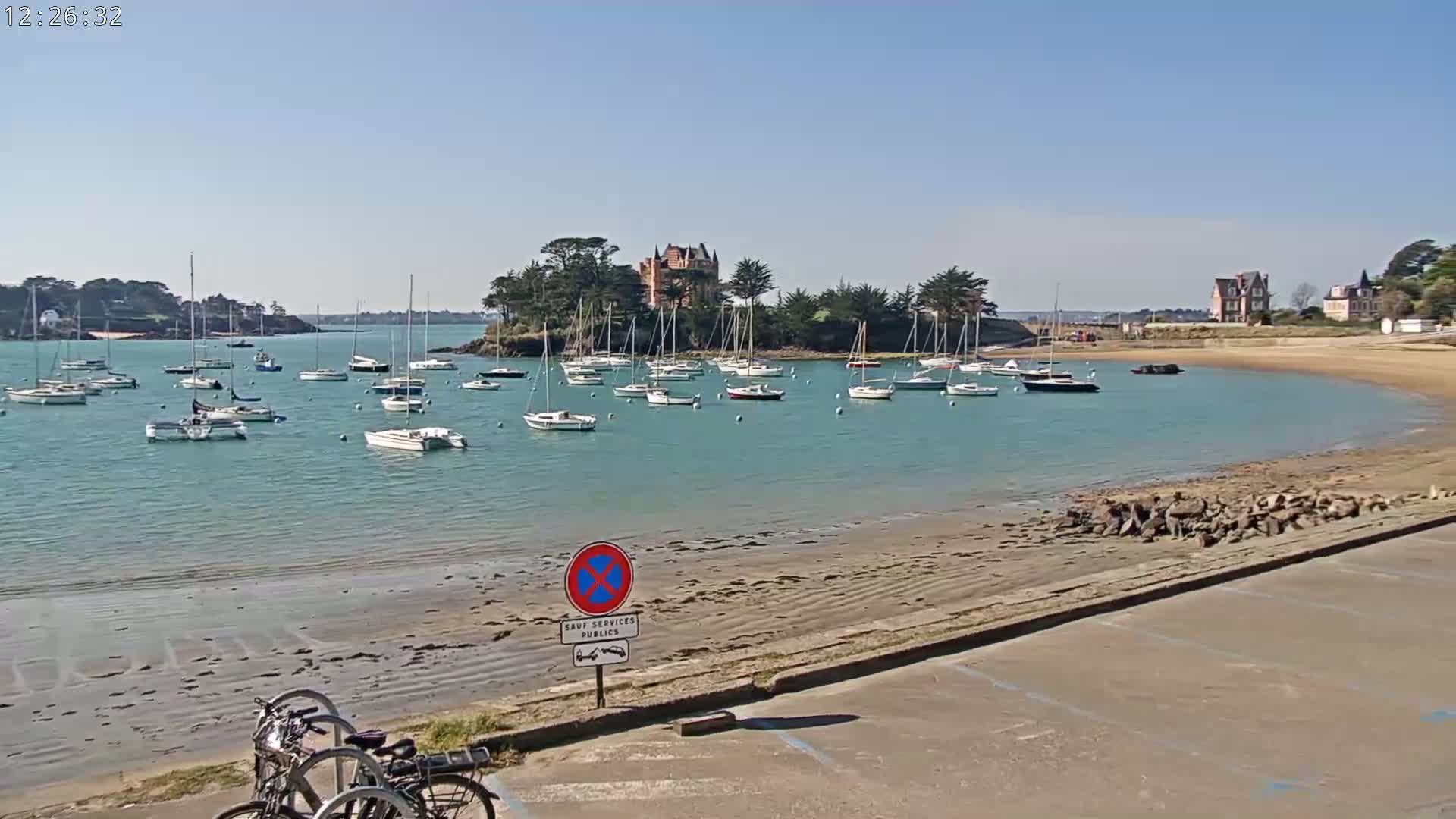 A calm, sunny day reveals a picturesque harbor filled with sailboats, a small island with a chateau, and a sandy shore with parked bicycles.