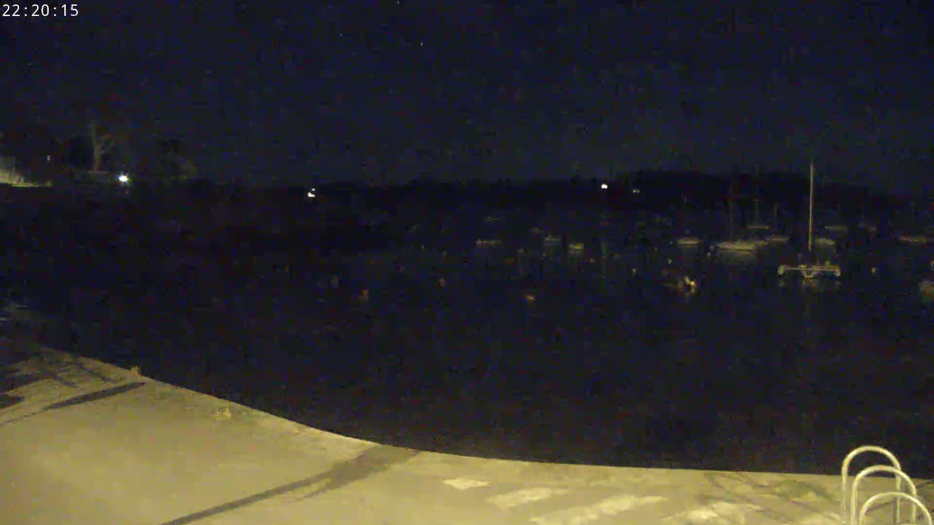 A dark body of water at night, partially visible under a clear, starry sky, is seen from a paved area with a ladder-like structure in the bottom right corner.