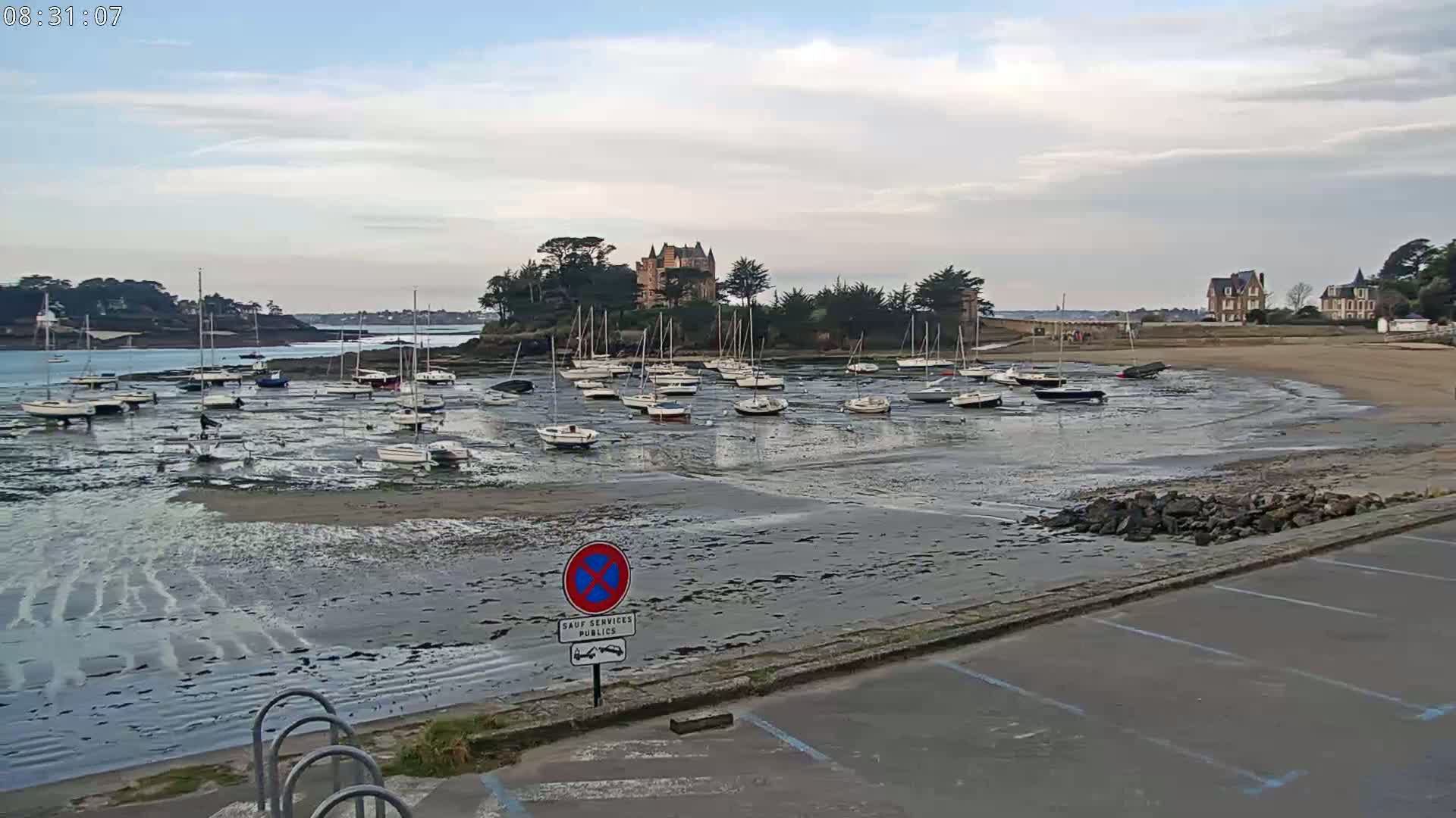 Many sailboats are grounded on a tidal flat near a château and other buildings on a cloudy day.
