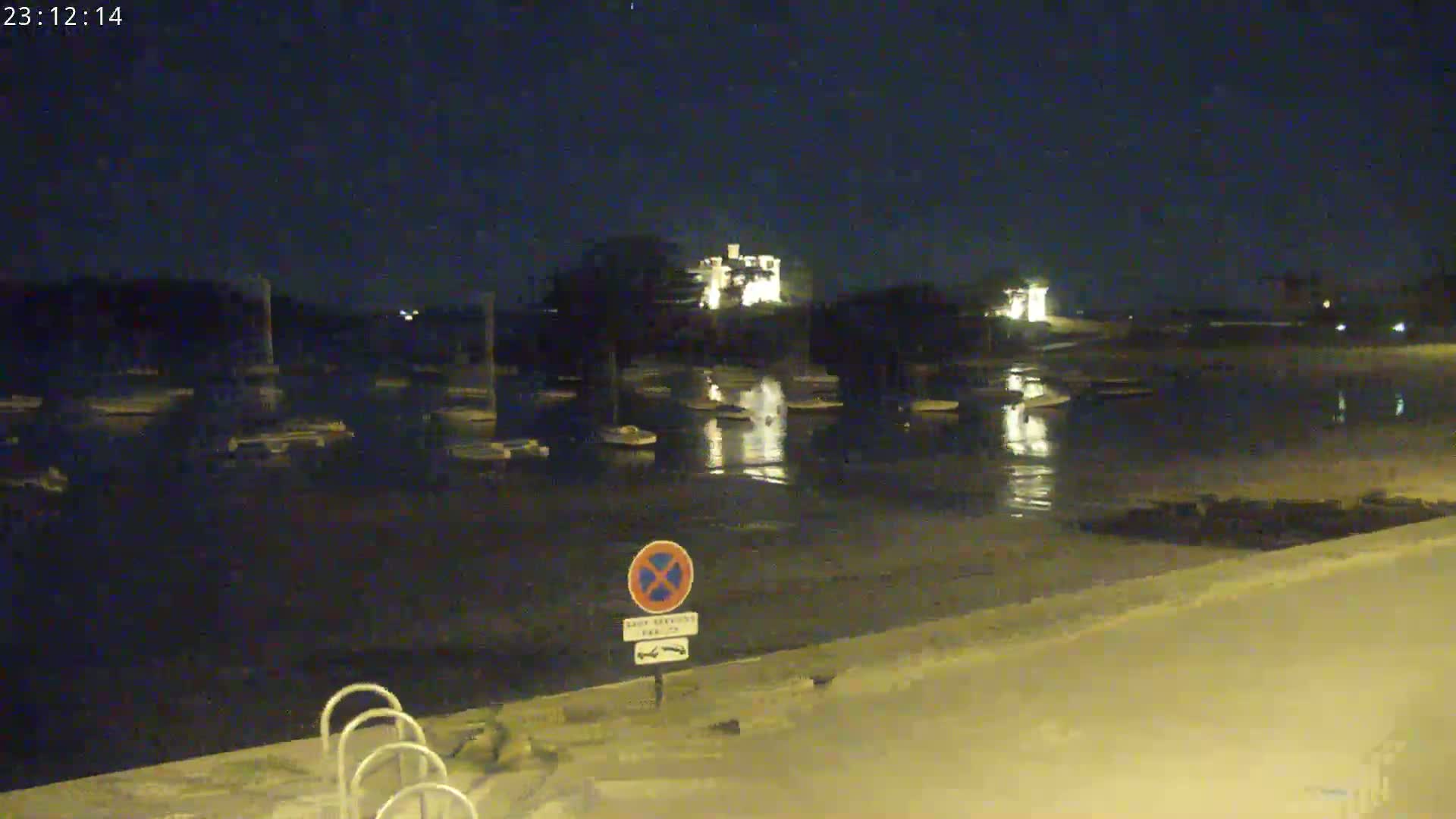 A nighttime view of a calm body of water with numerous boats moored, illuminated by lights from buildings on the far shore.