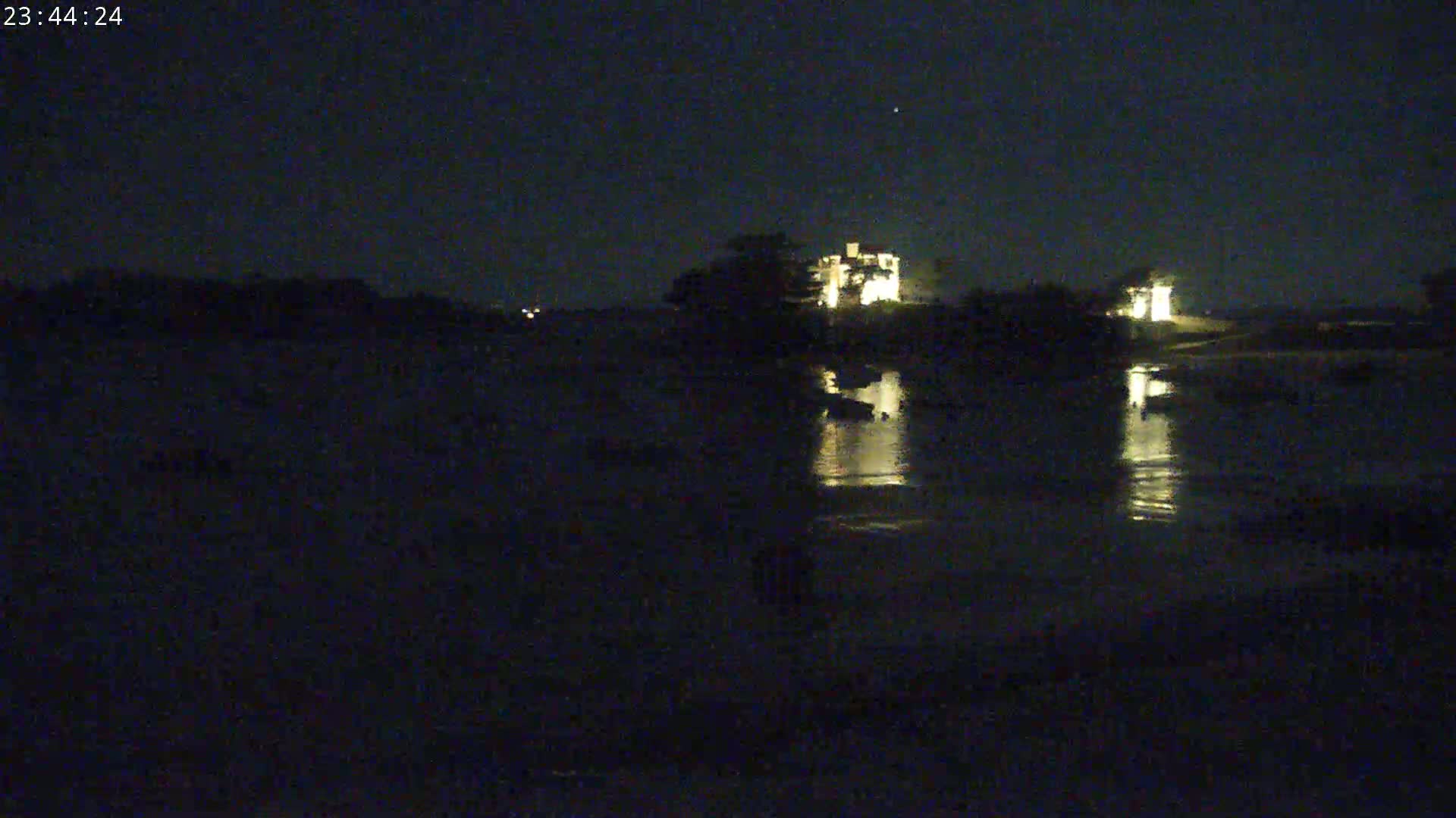 A nighttime view of a calm body of water reflecting the lights of a building on the opposite shore under a dark sky.