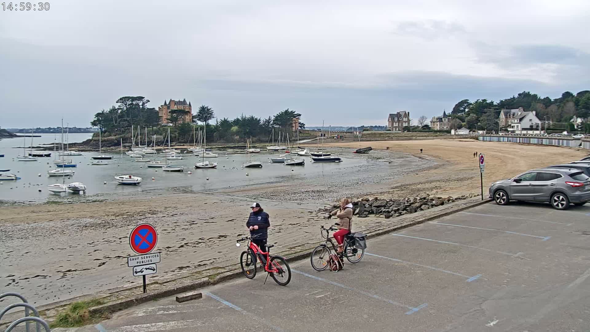 A sandy beach with numerous sailboats in the harbor, a few buildings on a small hill, two people standing with bicycles near a parking lot, all under an overcast sky.