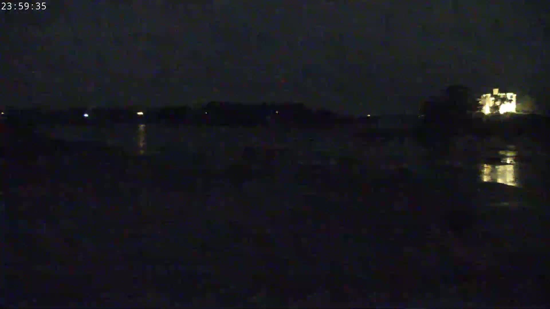 A dark, nighttime scene shows a body of water reflecting the lights of a building on the far shore under a mostly clear sky.