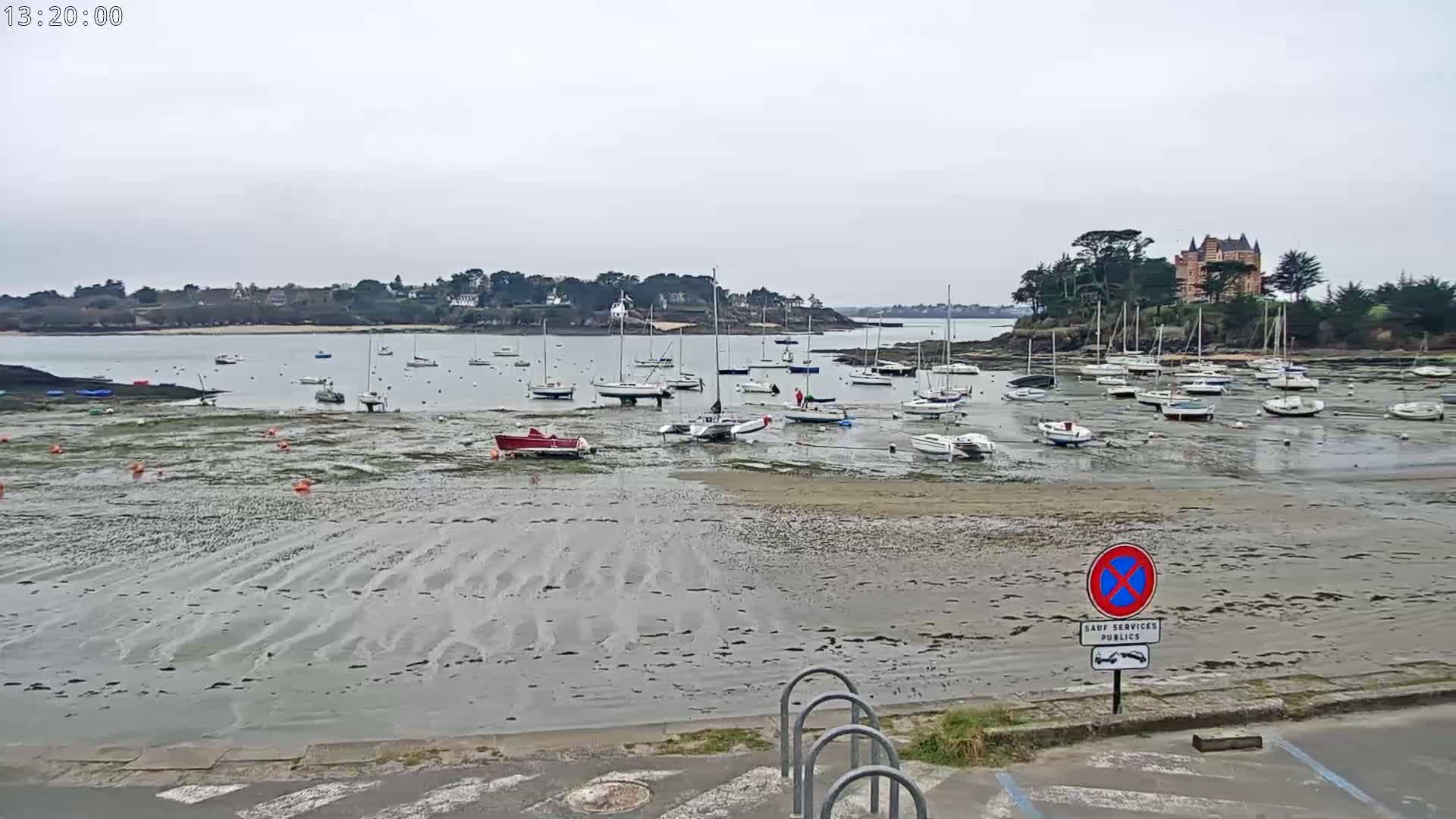 Numerous sailboats are grounded on a tidal flat near a shoreline with a chateau-like building visible in the background, under an overcast sky.