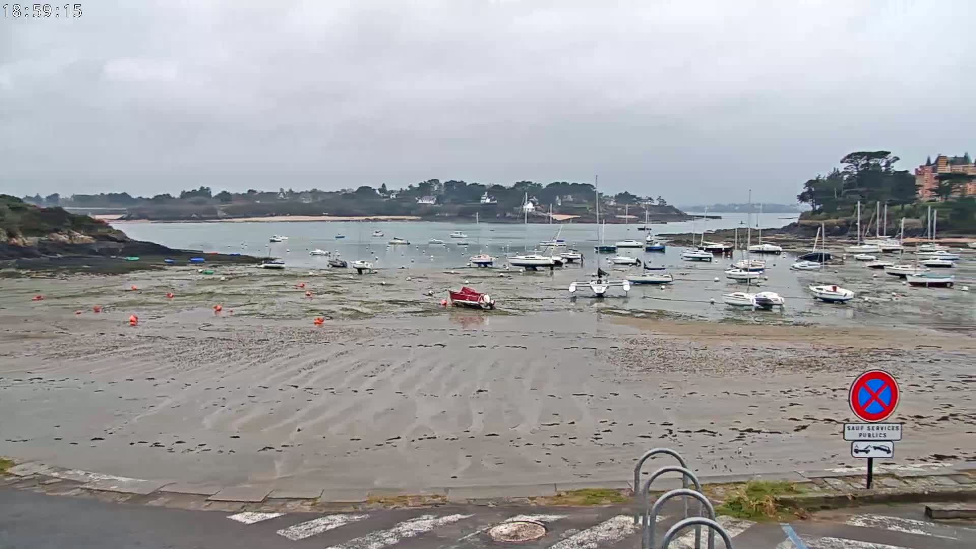 A mostly cloudy day reveals a tidal flat filled with numerous small boats at rest, some grounded in shallow water, with a calm bay and distant shoreline visible in the background.