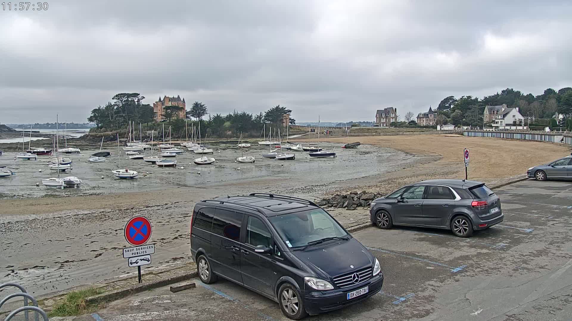 A sandy beach with numerous sailboats grounded at low tide, a château visible in the distance, and several cars parked nearby under an overcast sky.