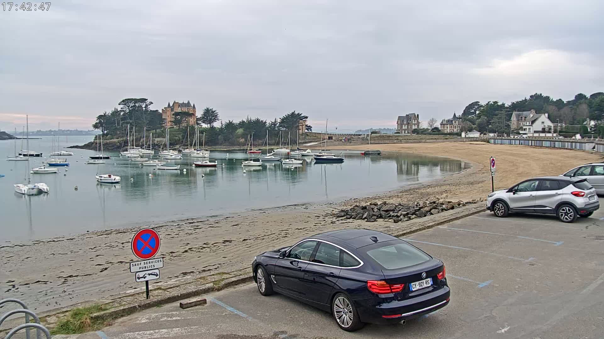 A calm bay with numerous sailboats is seen from a parking lot next to a sandy beach, with a few cars parked, under an overcast sky.
