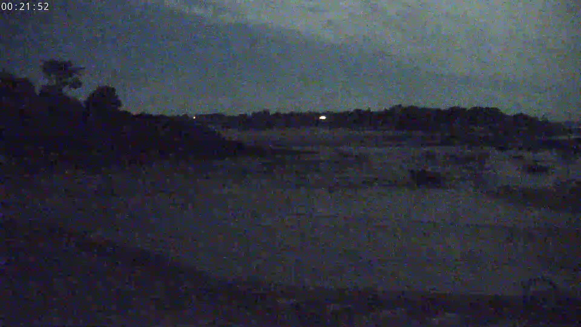 A dark, nighttime scene shows a flat, dark expanse of land with silhouetted trees and distant, faint lights under a dusky sky.