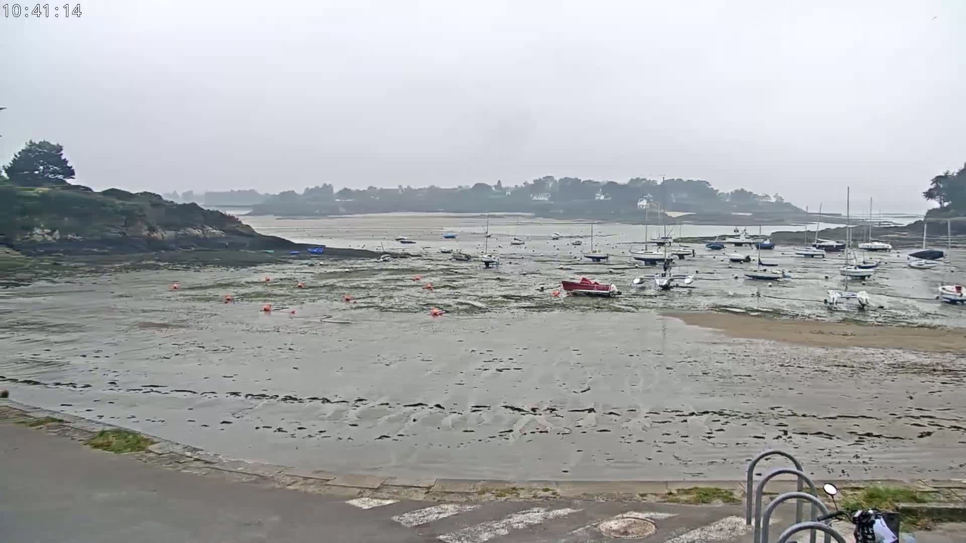 A mostly cloudy day reveals a tidal harbor with numerous small boats resting on exposed mudflats, surrounded by rocky shorelines and a distant tree-lined coast.