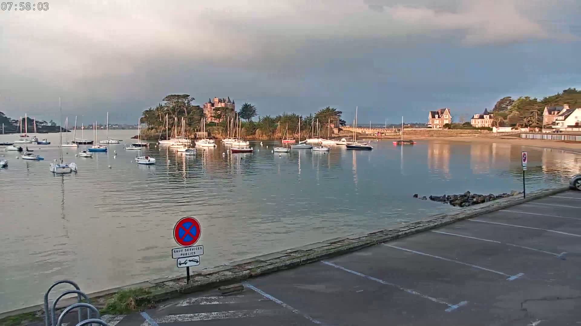 A calm harbor filled with sailboats is seen under a mostly cloudy sky, with buildings and a sandy shore in the background.