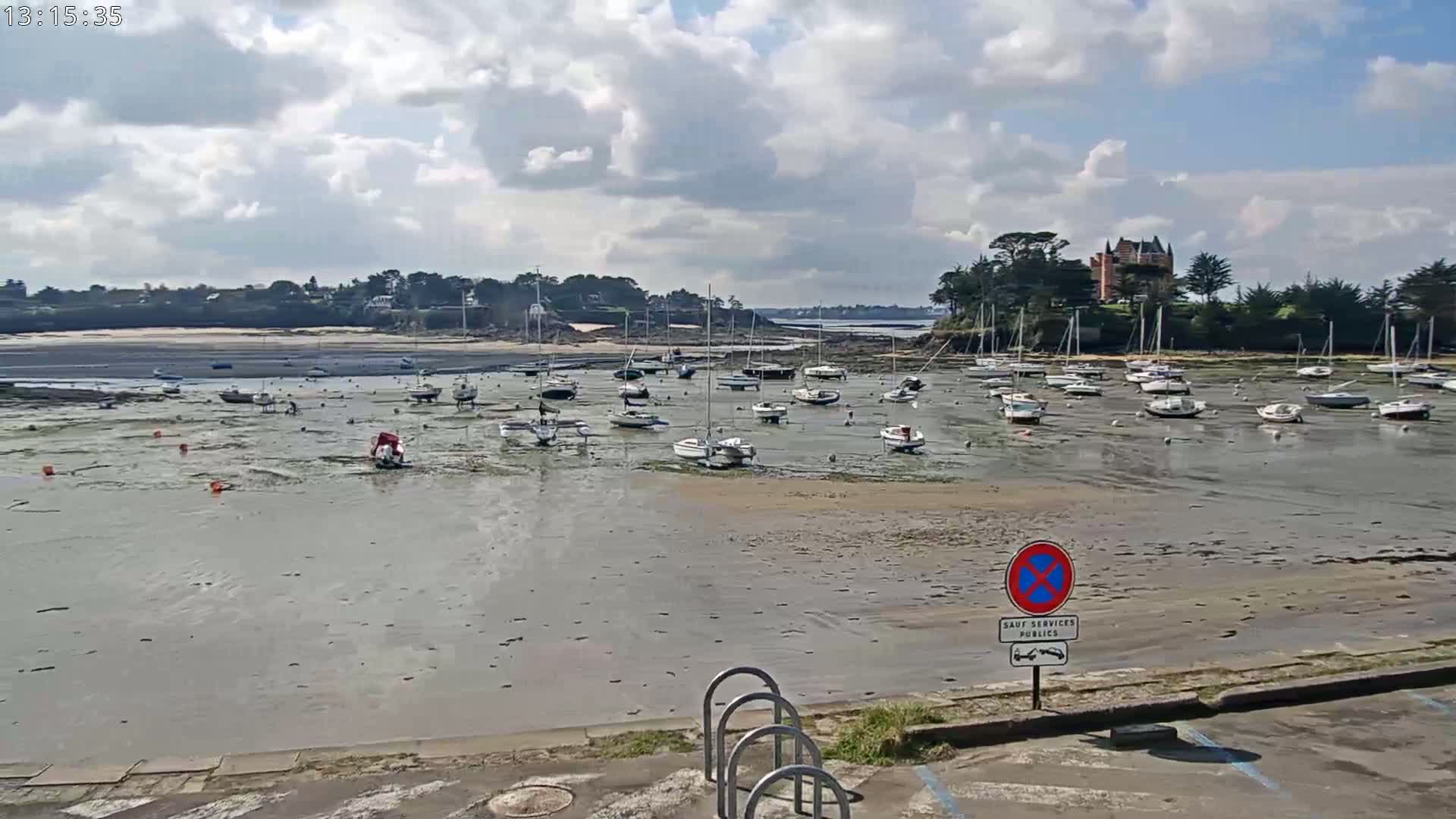 Numerous sailboats are grounded on a tidal flat, with a partly cloudy sky overhead.