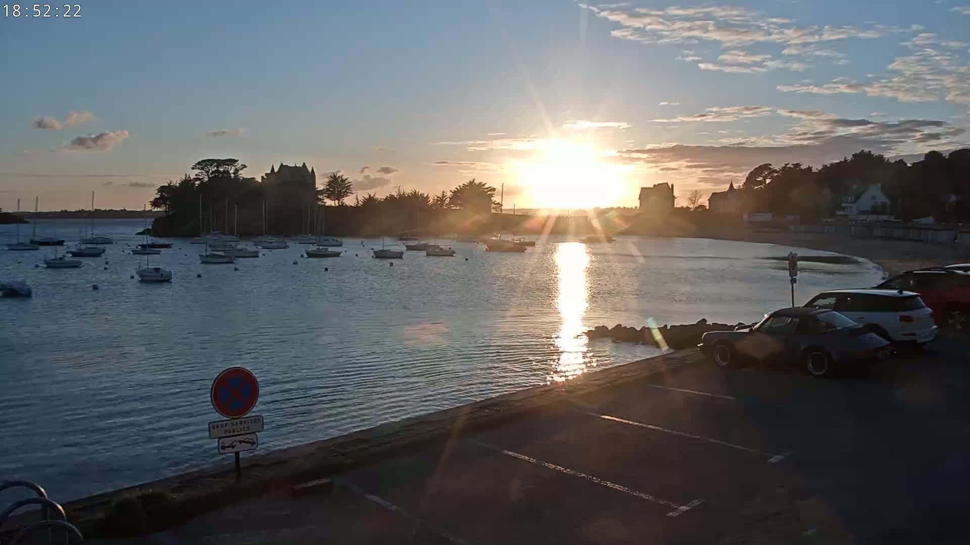 A calm bay with numerous sailboats is seen at sunset, with a stone building on a small peninsula and cars parked in a lot in the foreground.