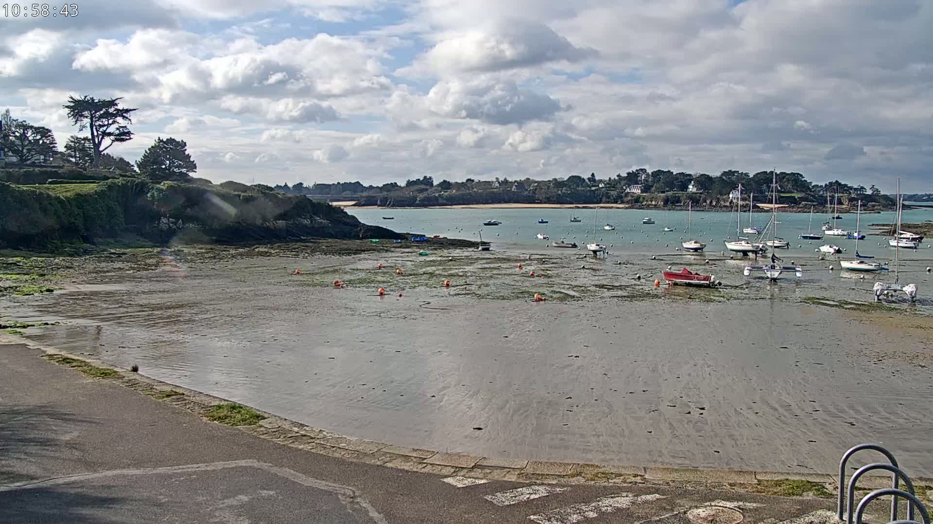 A mostly sunny day reveals a calm bay with numerous sailboats and small motorboats at low tide, anchored in shallow water near a rocky shoreline and green hills.