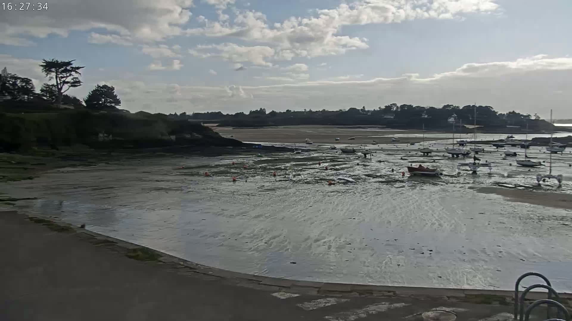 Under a partly cloudy sky, a calm body of water reveals a sandy bottom where numerous small boats are beached at low tide, with a shoreline of dark rocks and green vegetation visible in the distance.