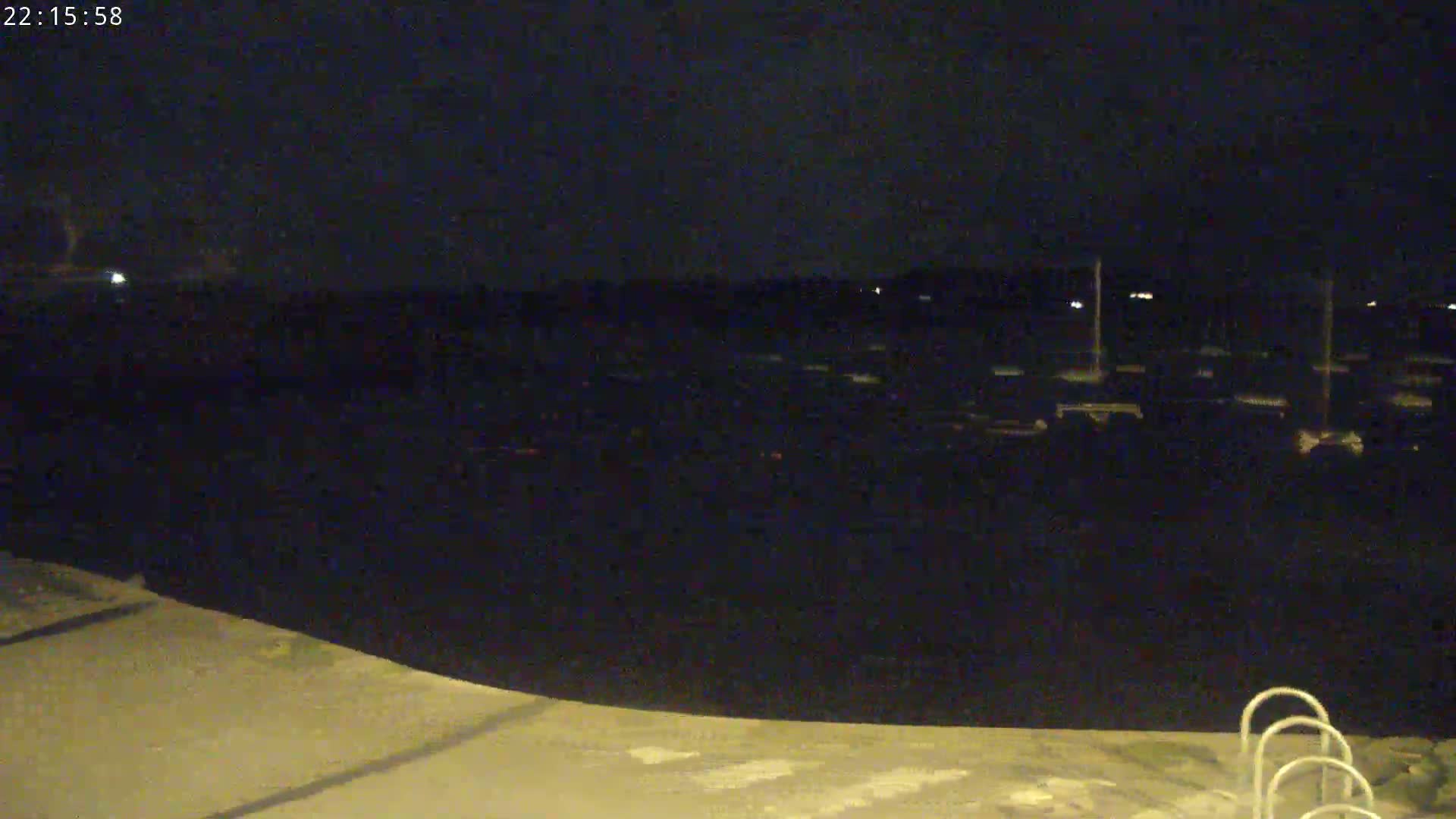 A nighttime view of a dark body of water with numerous small lights sparsely distributed across its surface, and a concrete area in the foreground featuring a portion of what appears to be pool ladder.