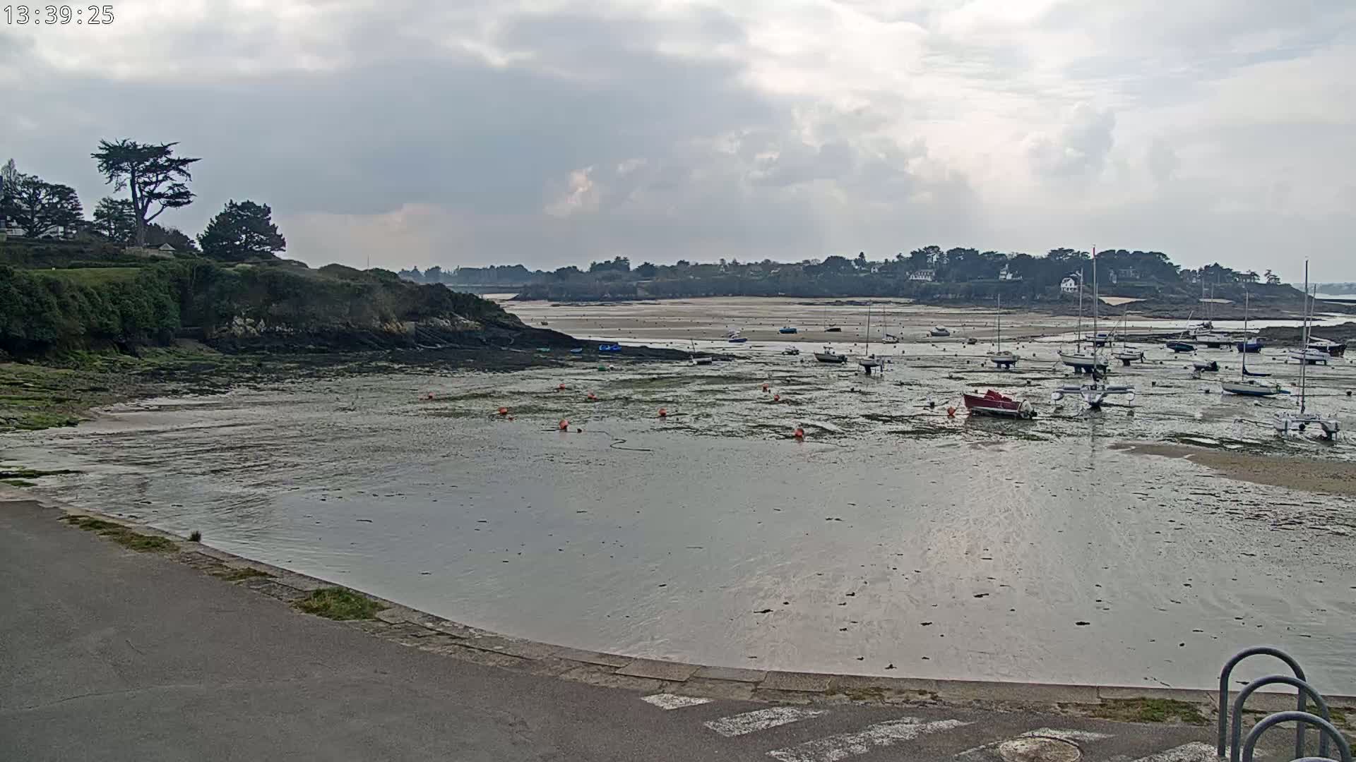A mostly cloudy day reveals a calm bay with numerous sailboats and small motorboats resting on exposed mudflats at low tide, nestled between green hillsides.