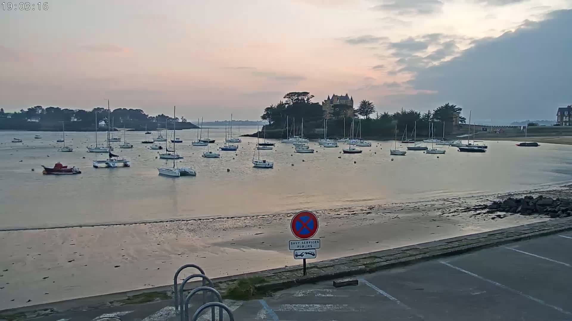 Many sailboats are moored in a calm bay under a cloudy, pinkish sunset sky, near a shoreline with a small chateau on an island.